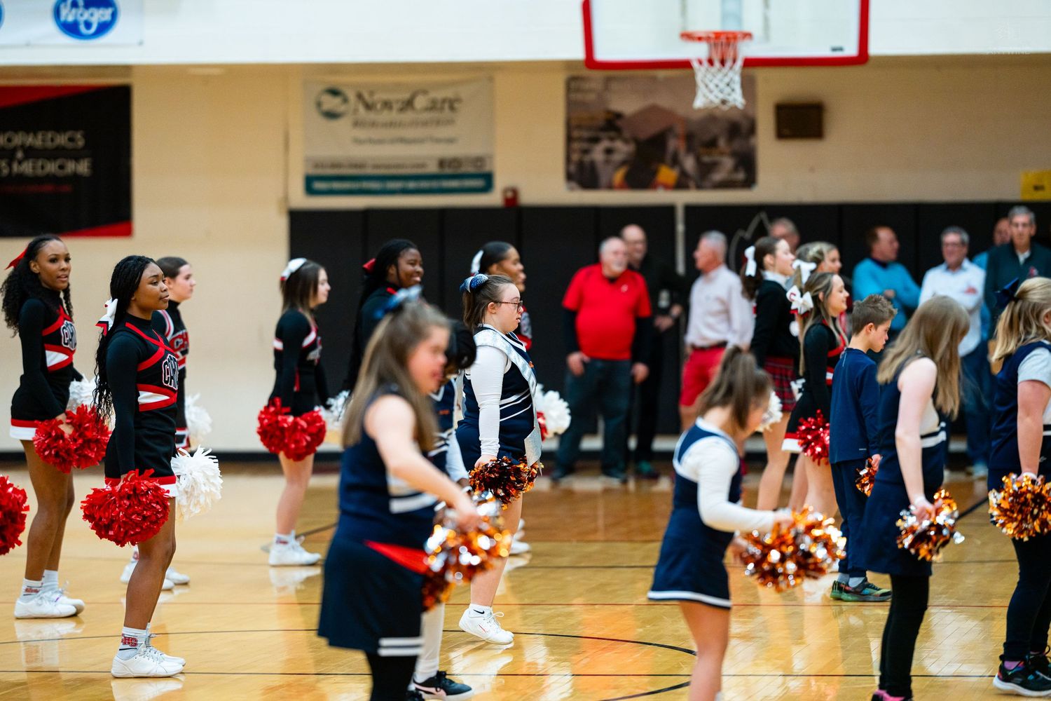 Cheerleaders in red and navy uniforms perform with colorful pom-poms in a gymnasium with sponsors on walls.