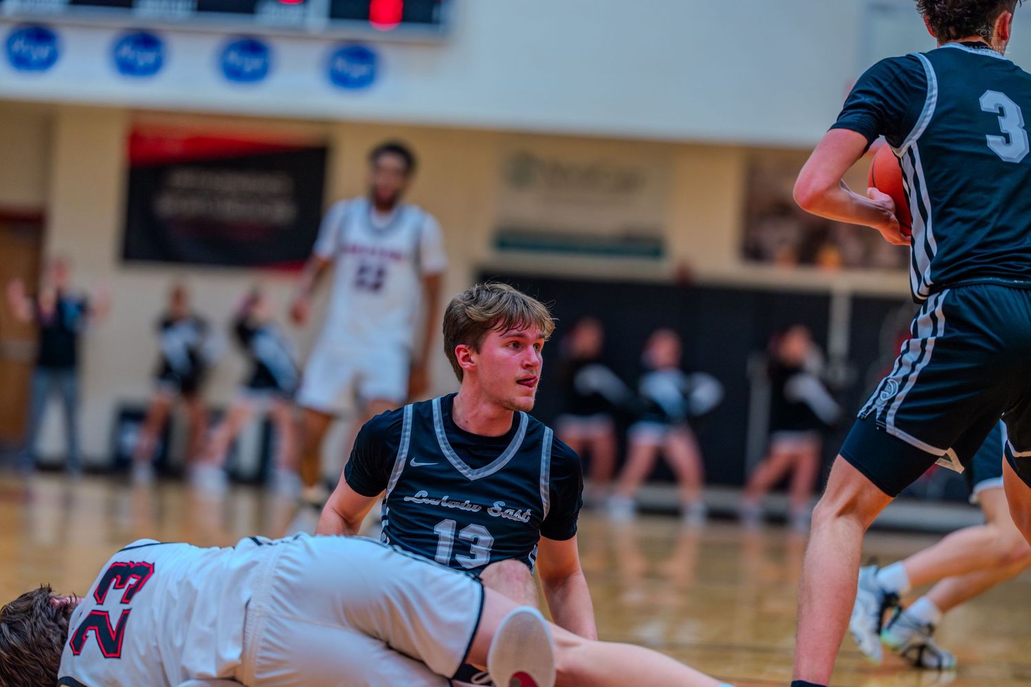 Player #13 in dark uniform looks down at a fallen player during a high school basketball game.