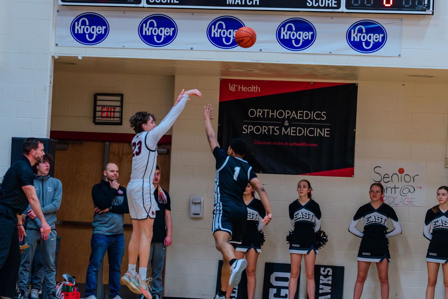 A basketball player attempts a shot near the sideline during a high school game with cheerleaders watching nearby.
