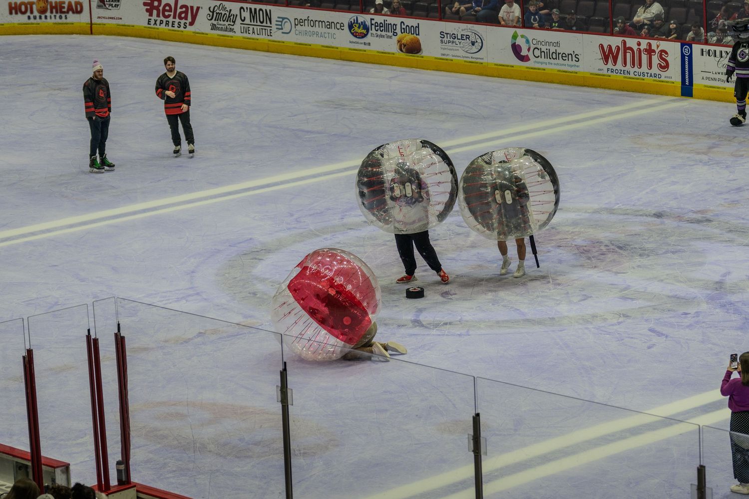 Participants in transparent and red inflatable zorb balls play bubble soccer on an indoor ice rink.