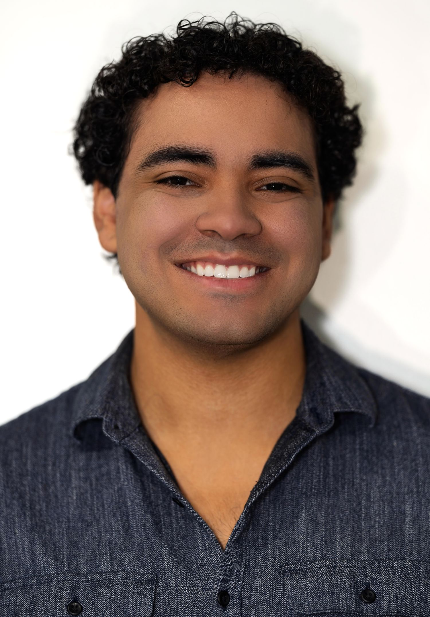 A smiling young man with curly dark hair wearing a gray collared shirt poses in a bright studio portrait.