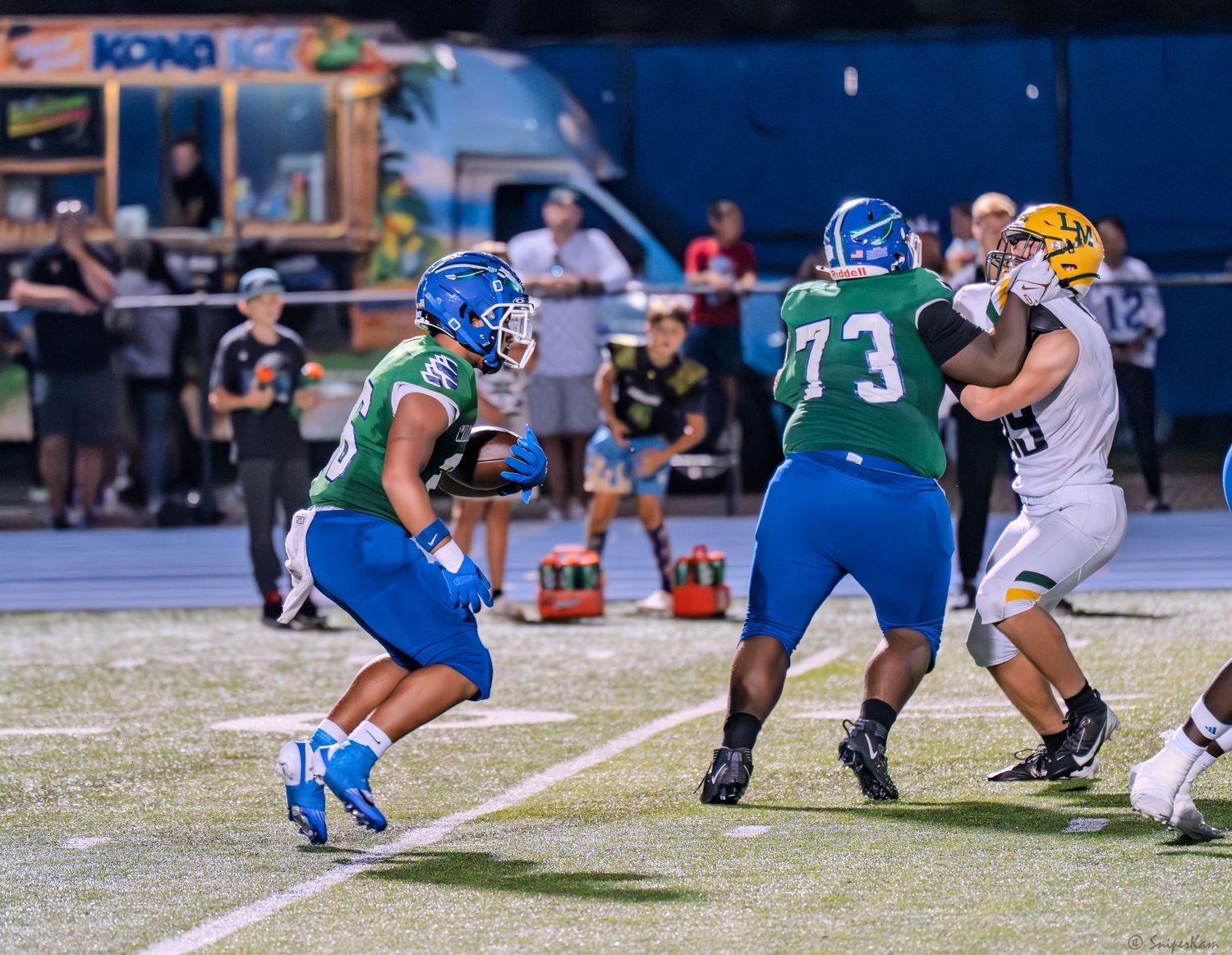 Warriors offensive lineman number 73 in green blocks for his quarterback during a high school football game.