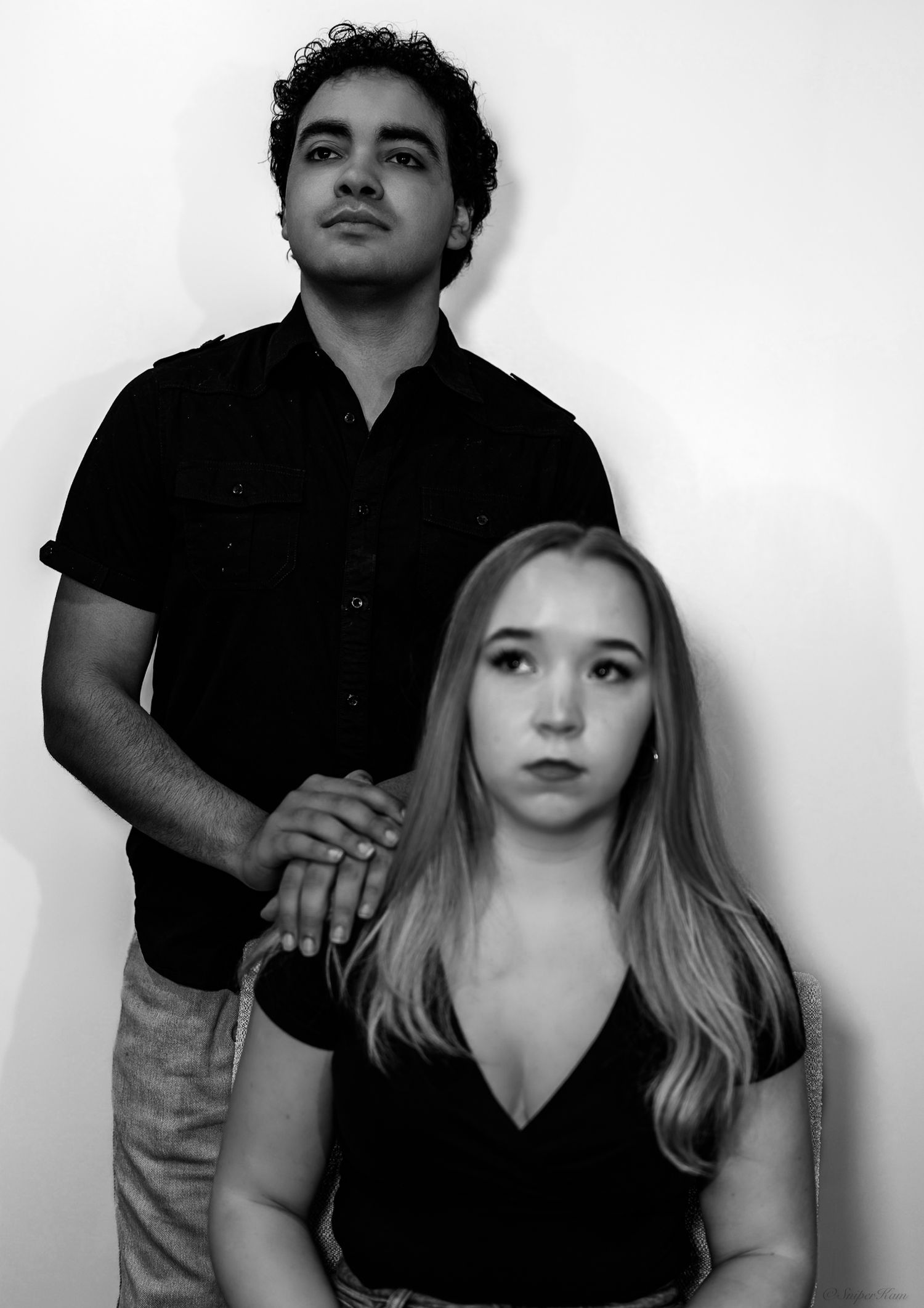 Black and white portrait of serious couple, man standing behind seated woman against white wall.