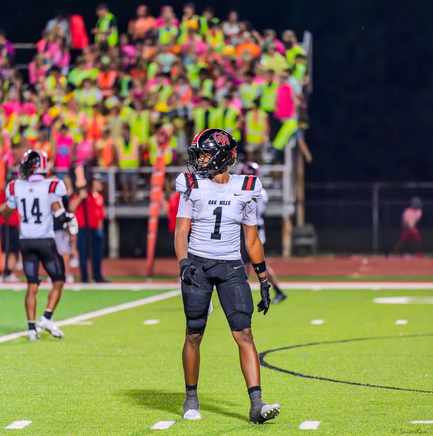 Football player #1 in dark uniform stands on field with colorful crowd visible in background.