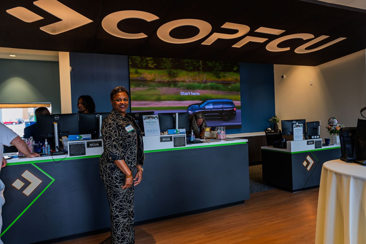 A smiling woman stands at the COPFCU credit union branch counter with computer workstations and green accents.