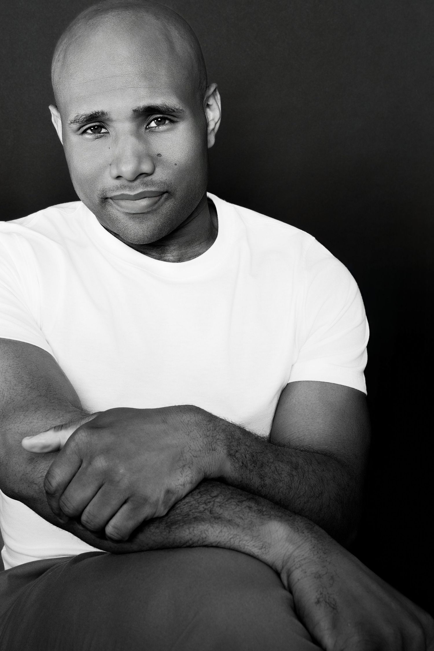 Black and white portrait of bald man in white t-shirt sitting relaxed against dark background.