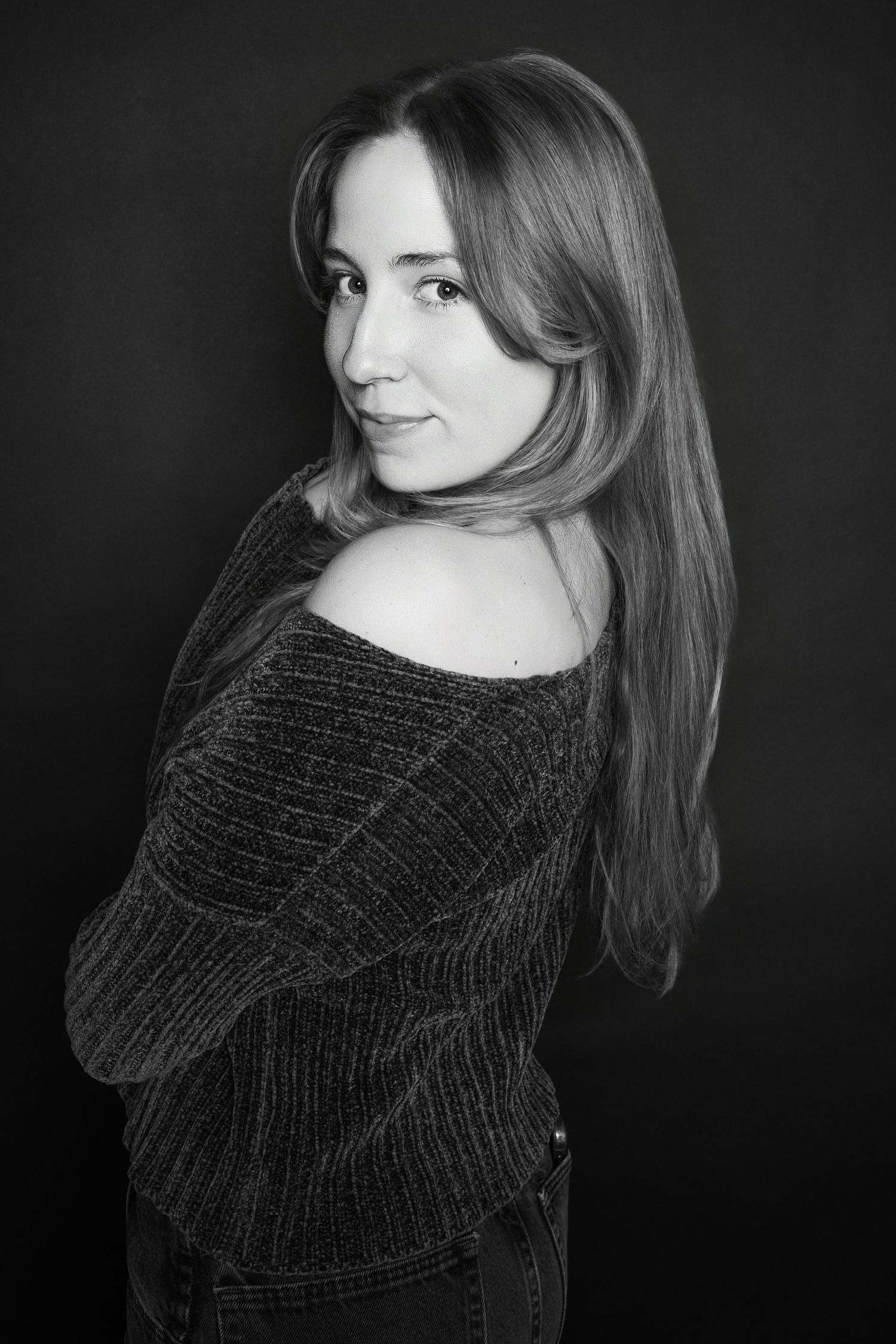Black and white portrait of woman in off-shoulder sweater posing against dark background.