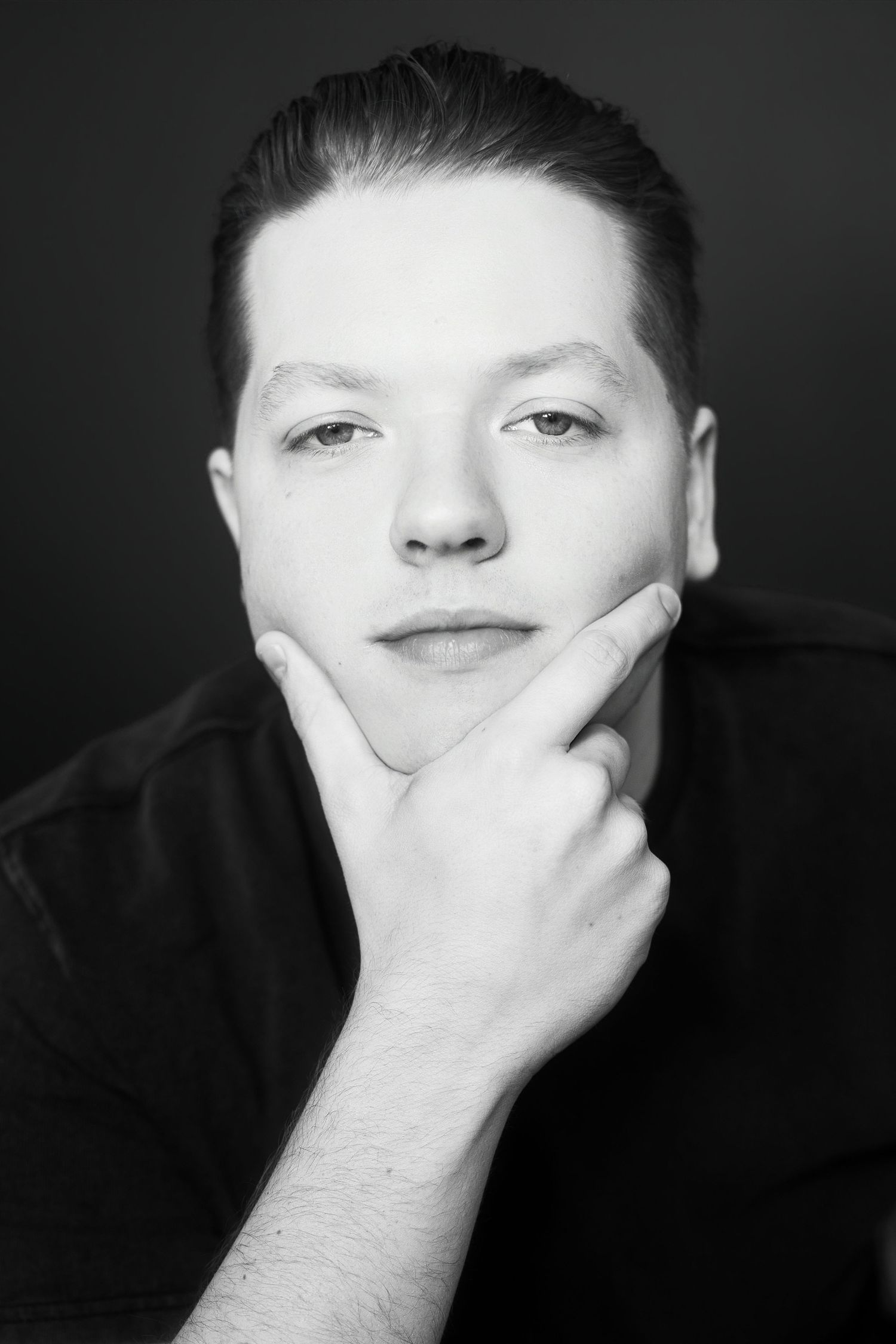 Black and white portrait of young man resting chin on hand against dark background.