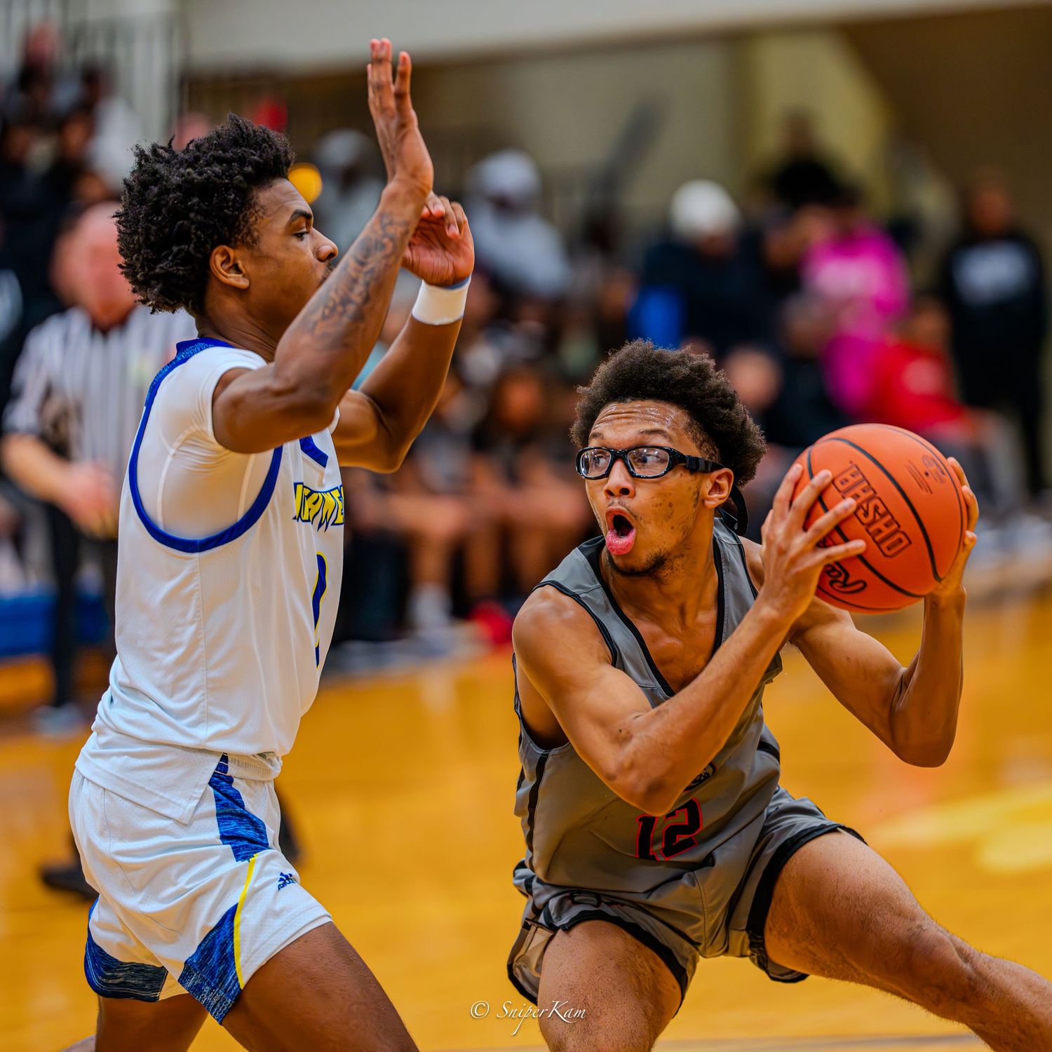 Basketball player in gray jersey drives to basket against defender in blue uniform.