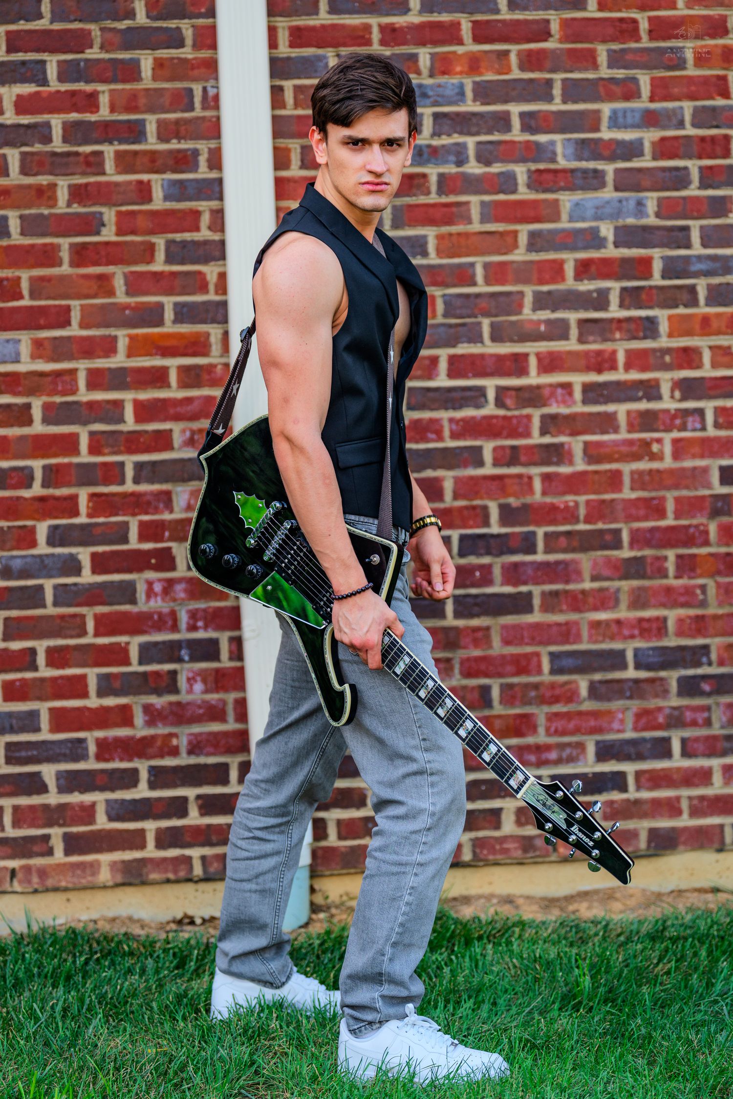 Young muscular man in sleeveless black vest holding electric guitar against red brick wall.