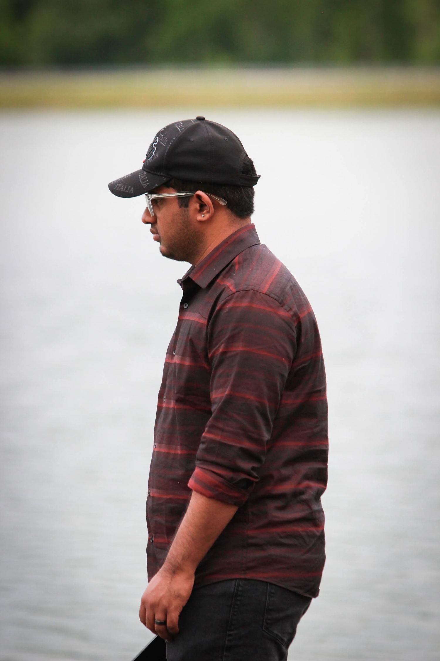 A heavyset man in a dark red striped shirt and black baseball cap stands in profile near a calm lake.