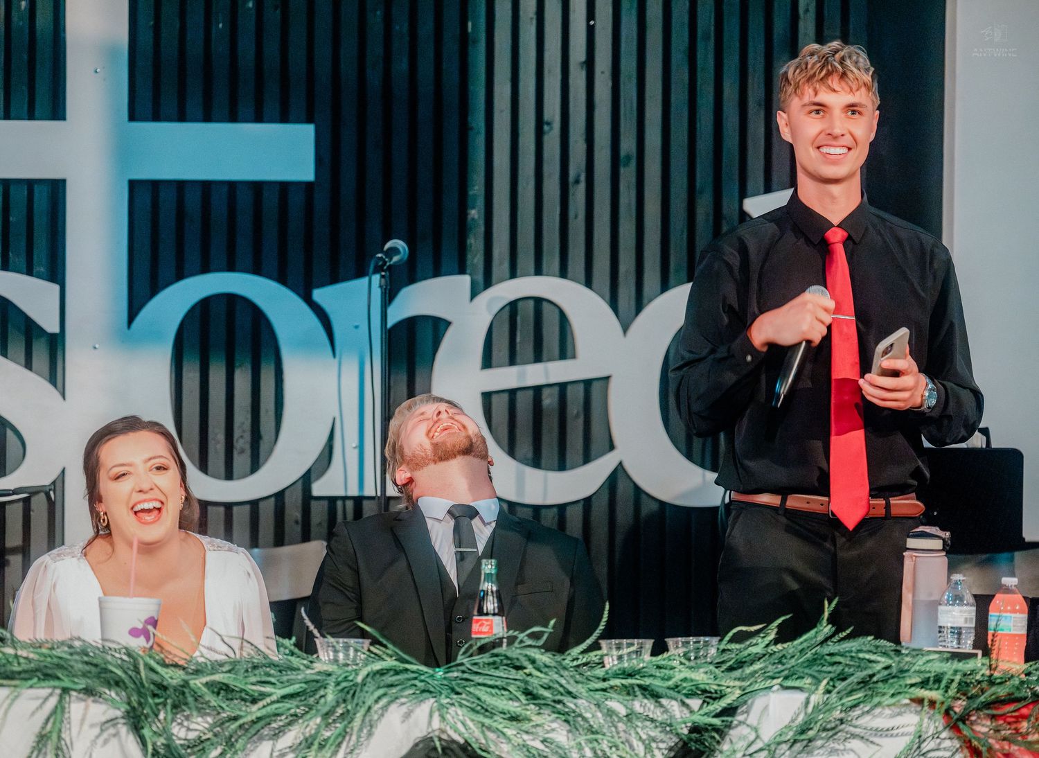 A groomsman in black suit and red tie delivers a speech as the bride and groom laugh at reception.