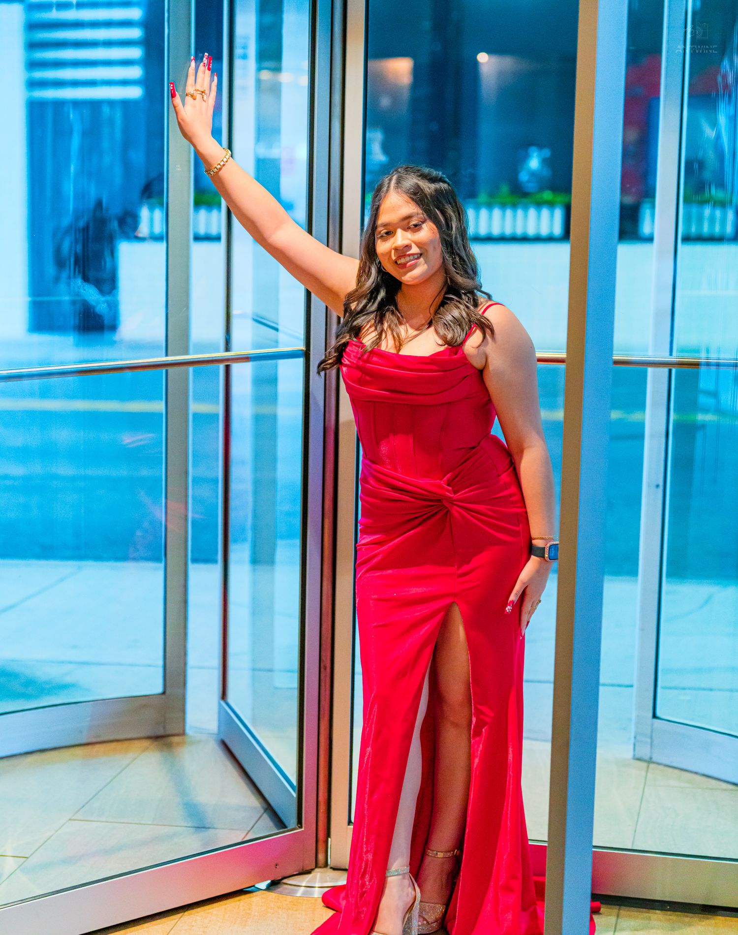 Woman in elegant red velvet slit gown waving at a formal venue entrance.