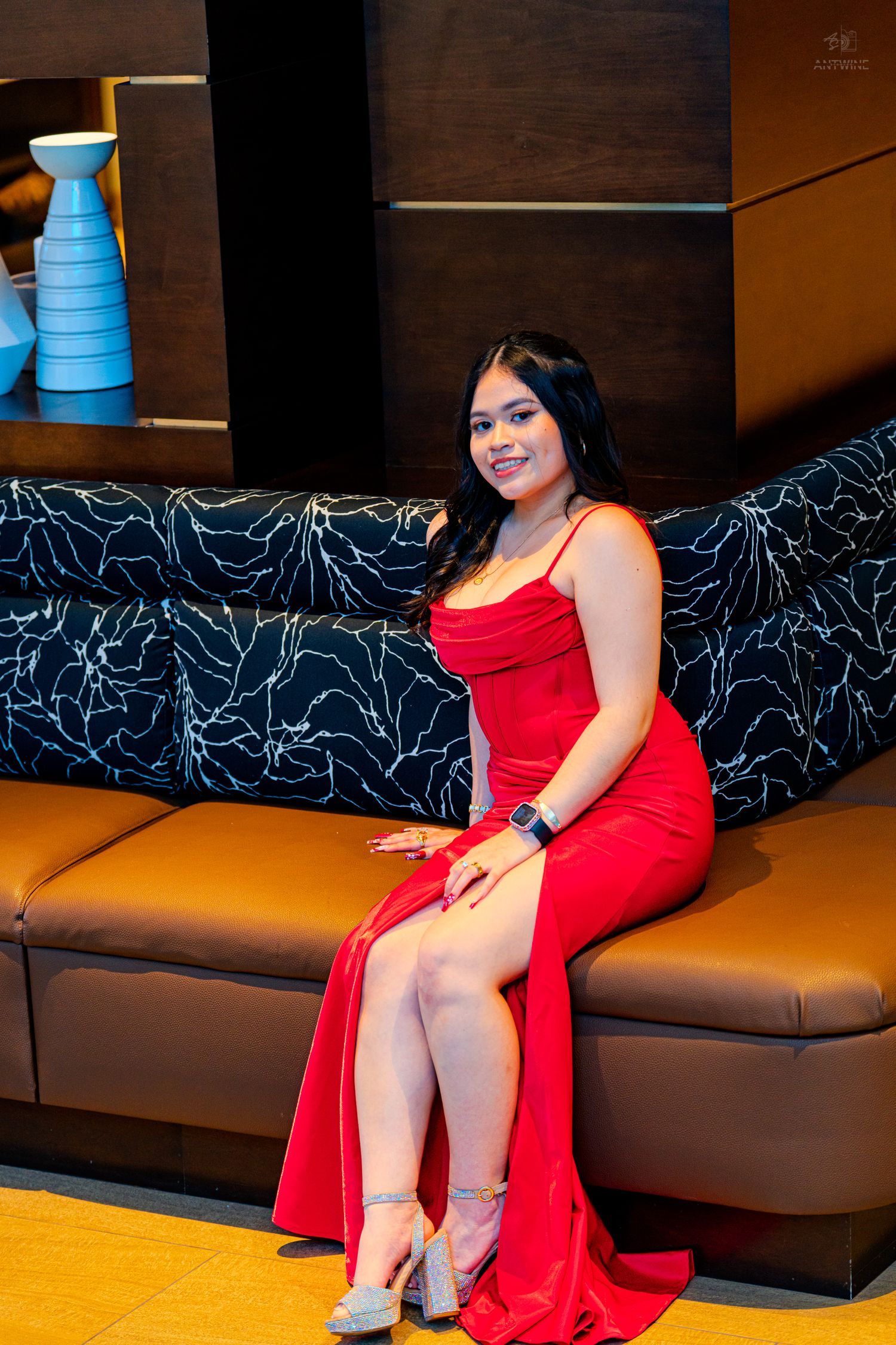 Young woman in elegant red slit gown and heels sitting on leather sofa smiling at camera.