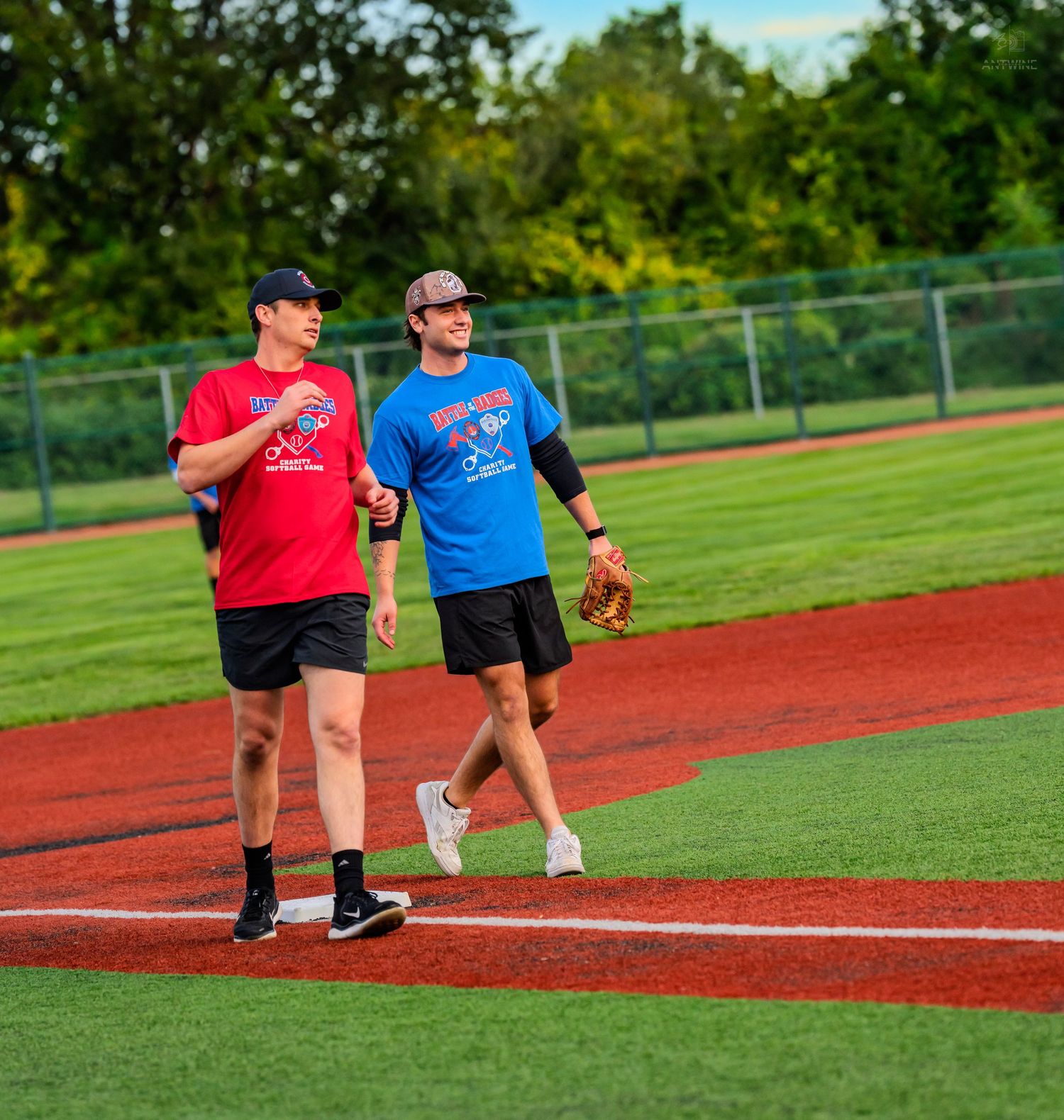 Two softball players in red and blue shirts walk together on a bright green baseball field.