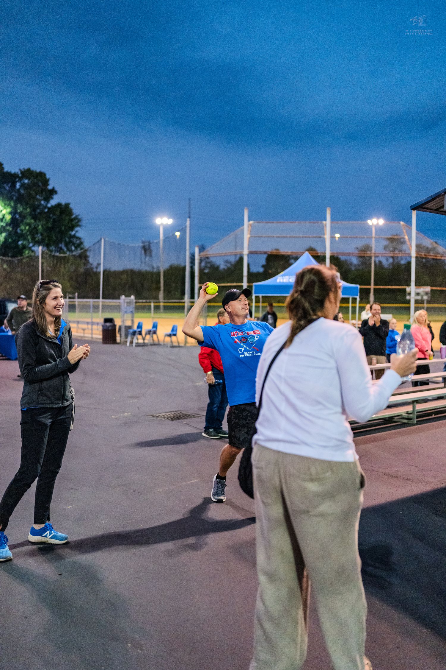 Man tosses a yellow softball to women during a nighttime recreational baseball practice under stadium lights.