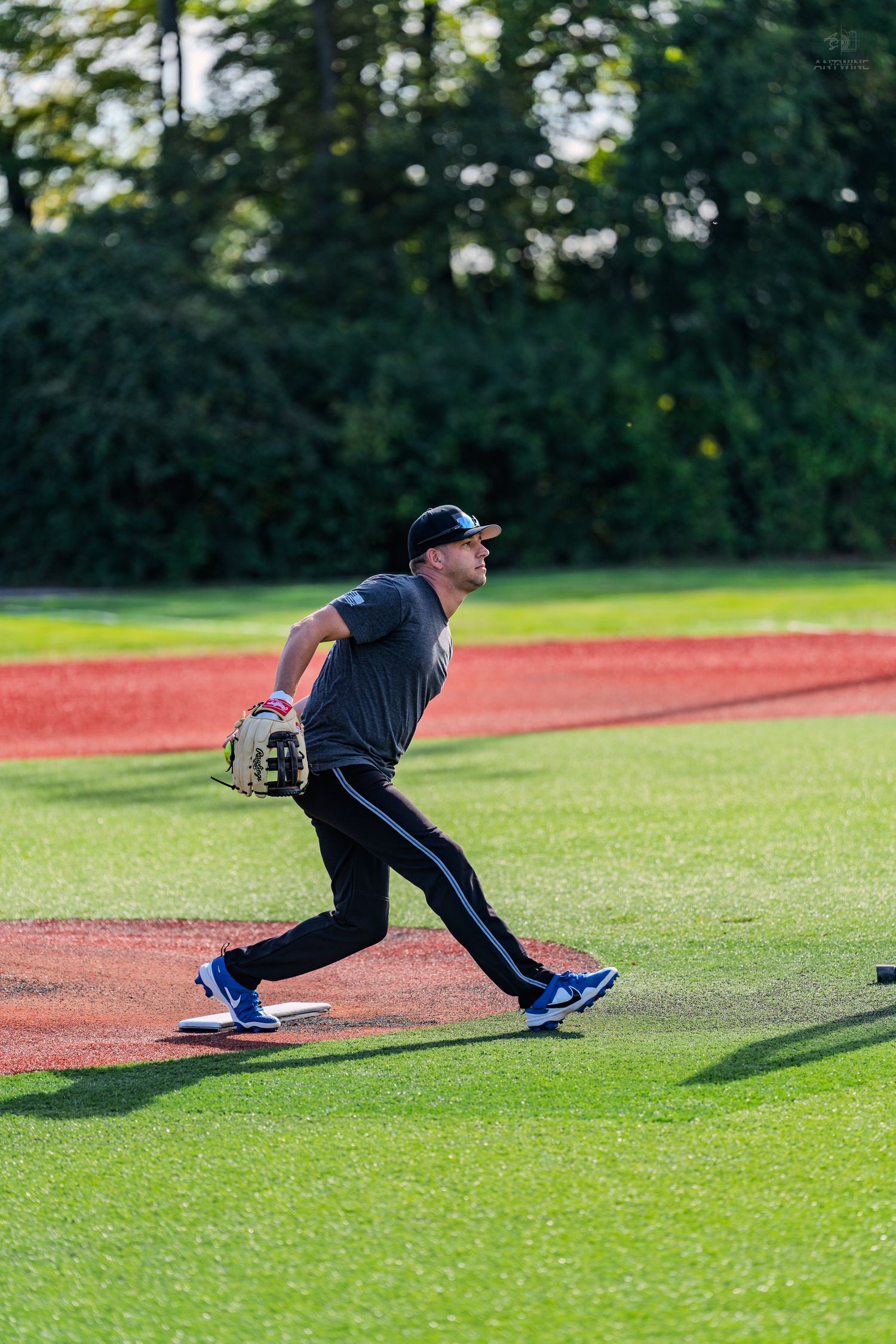 Baseball player in black uniform throwing ball on green field with red warning track and trees in background.