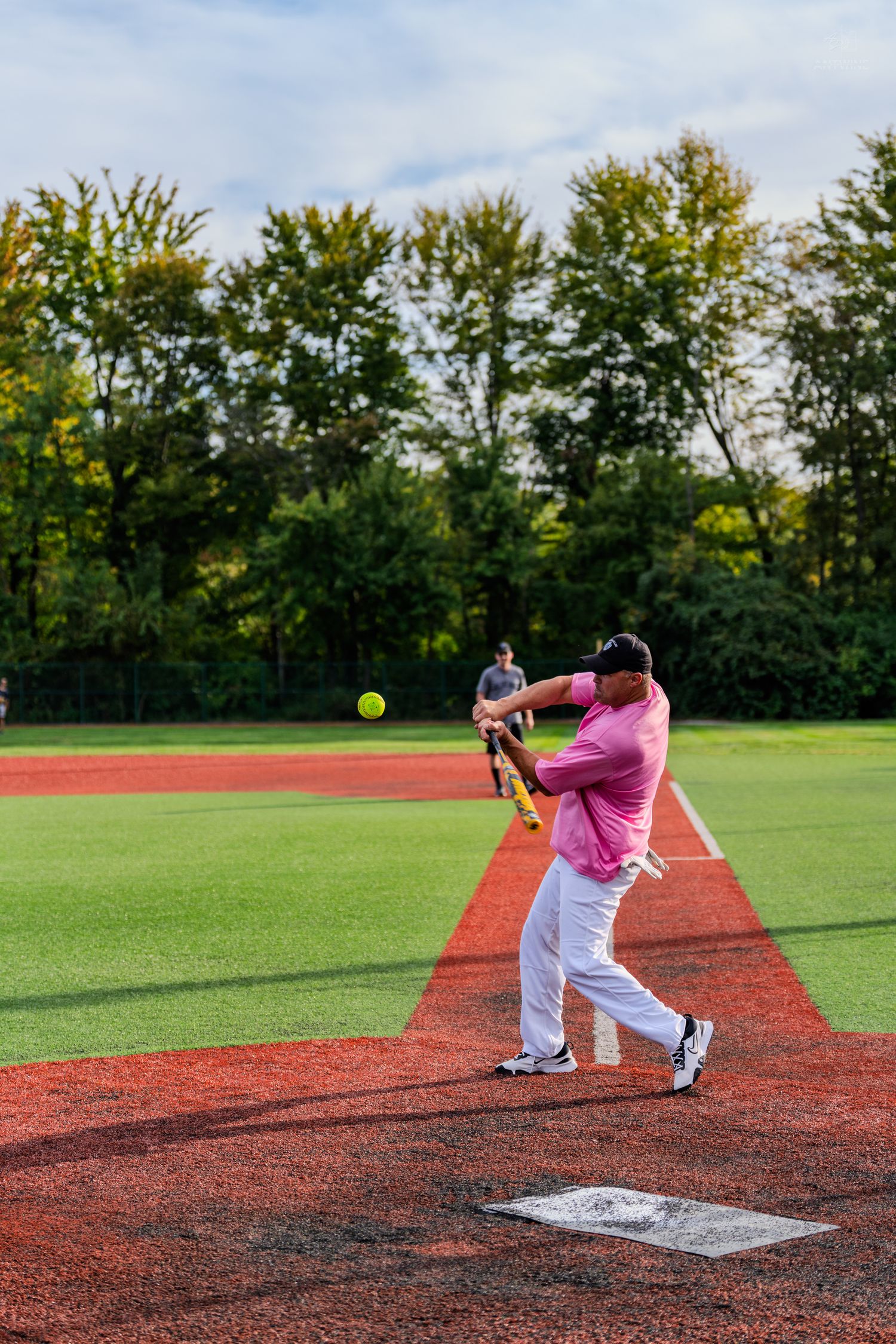 Batter in pink shirt swinging at a yellow softball at home plate on a well-maintained baseball field.
