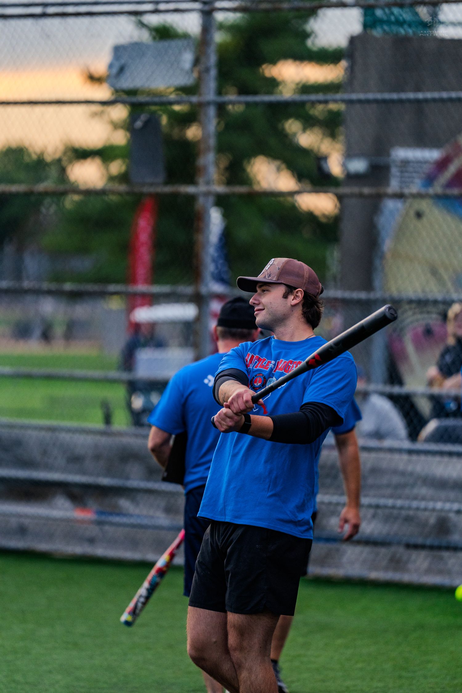 Smiling softball player in blue jersey holding a bat on a recreational field during an evening game.