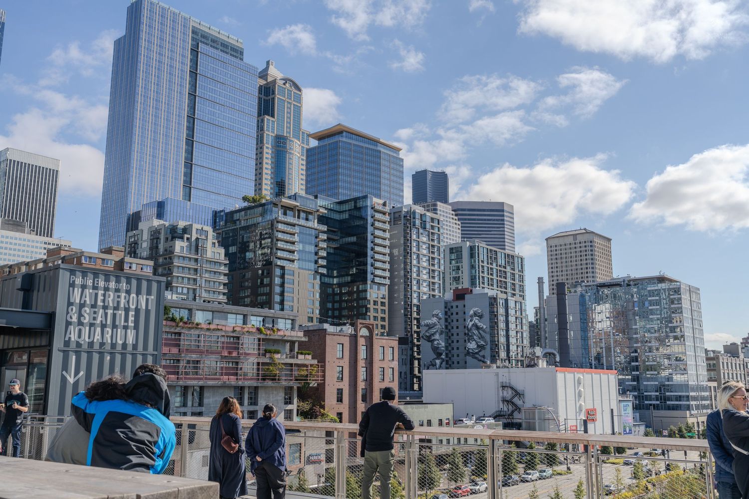 Tourists on a rooftop terrace overlooking Seattle's downtown skyline with modern skyscrapers under a partly cloudy sky.
