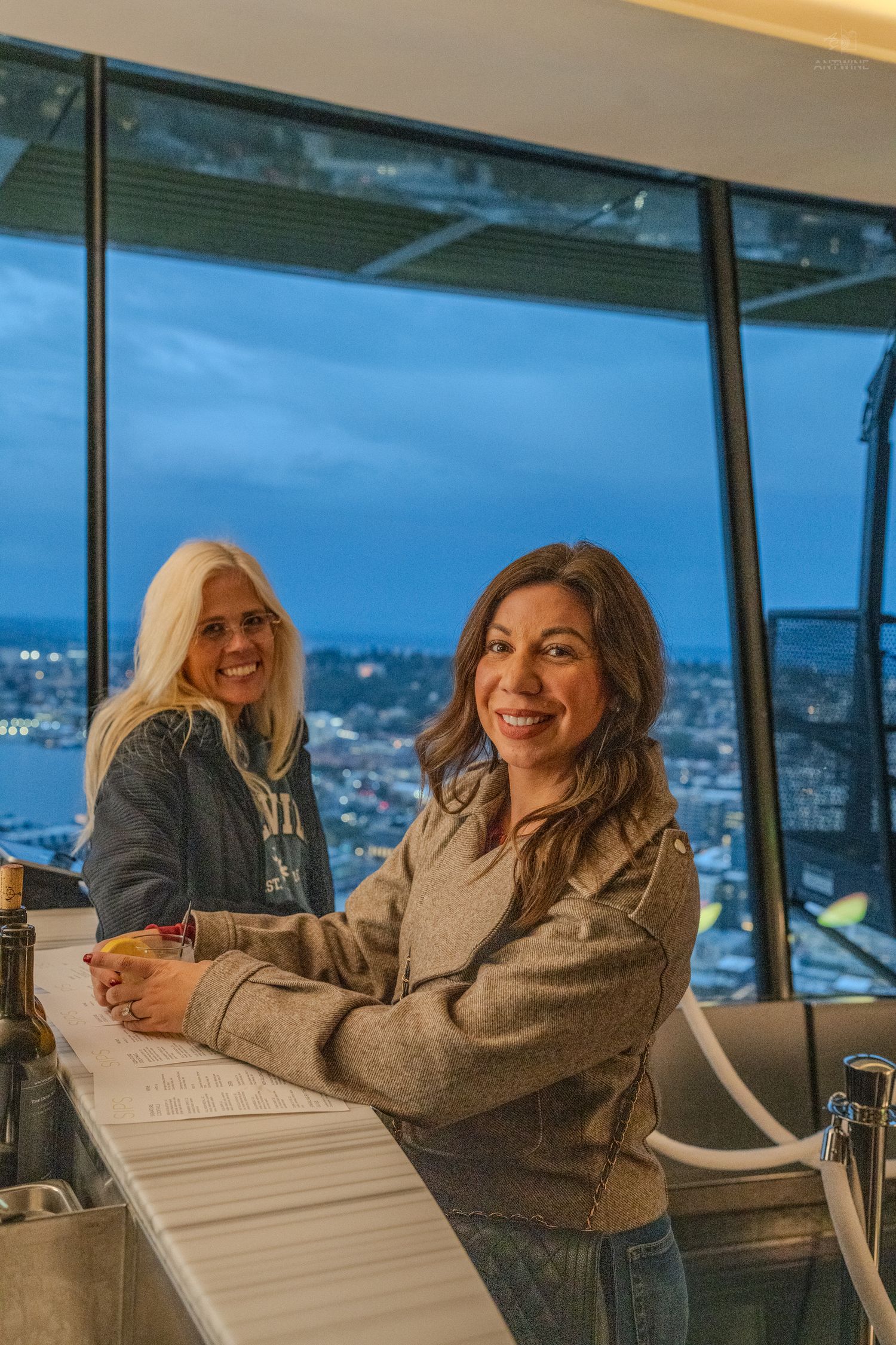 Two women smile at a high-rise bar with a stunning city skyline view at dusk.