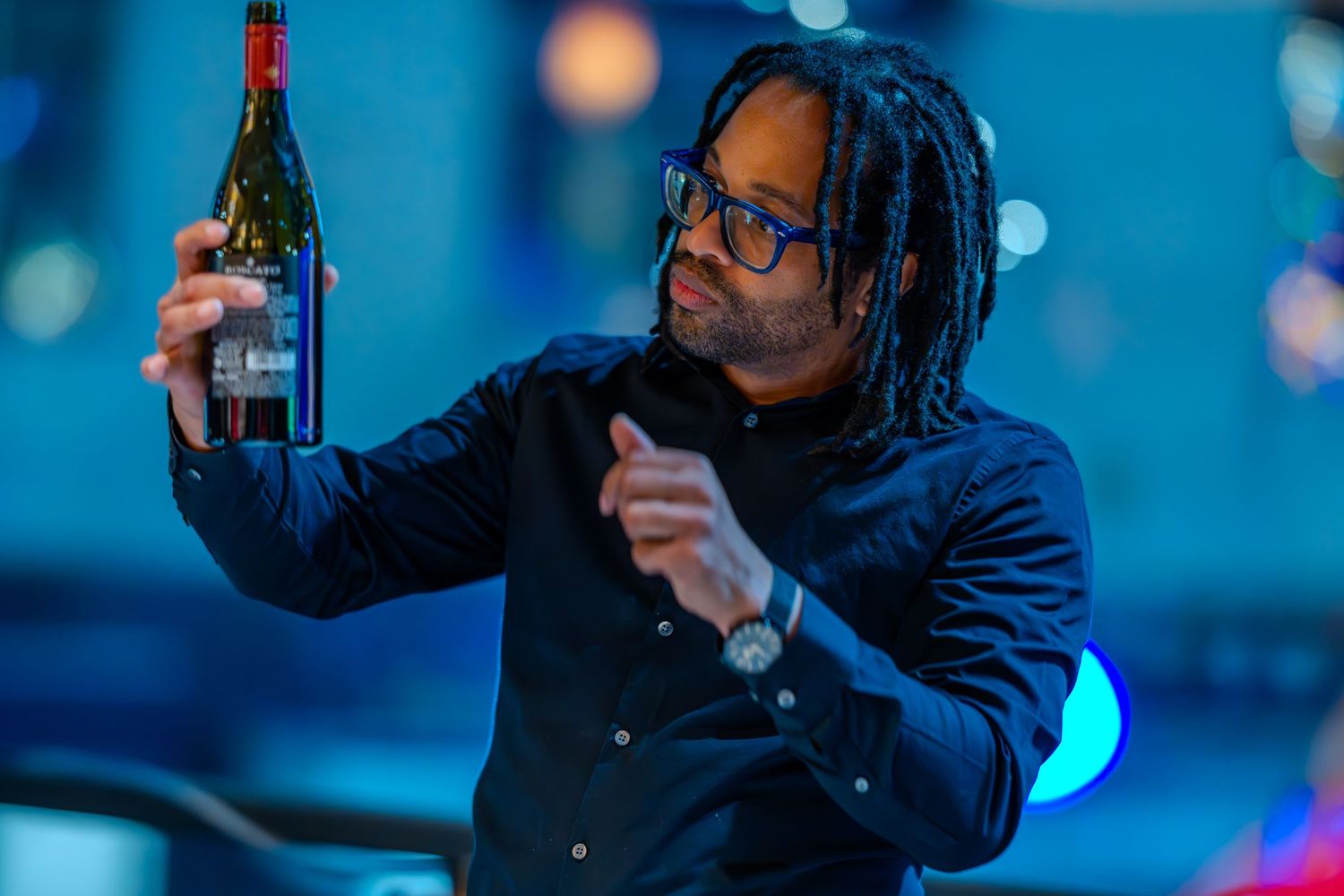 Man with dreadlocks and glasses examining a red wine bottle outdoors at night.