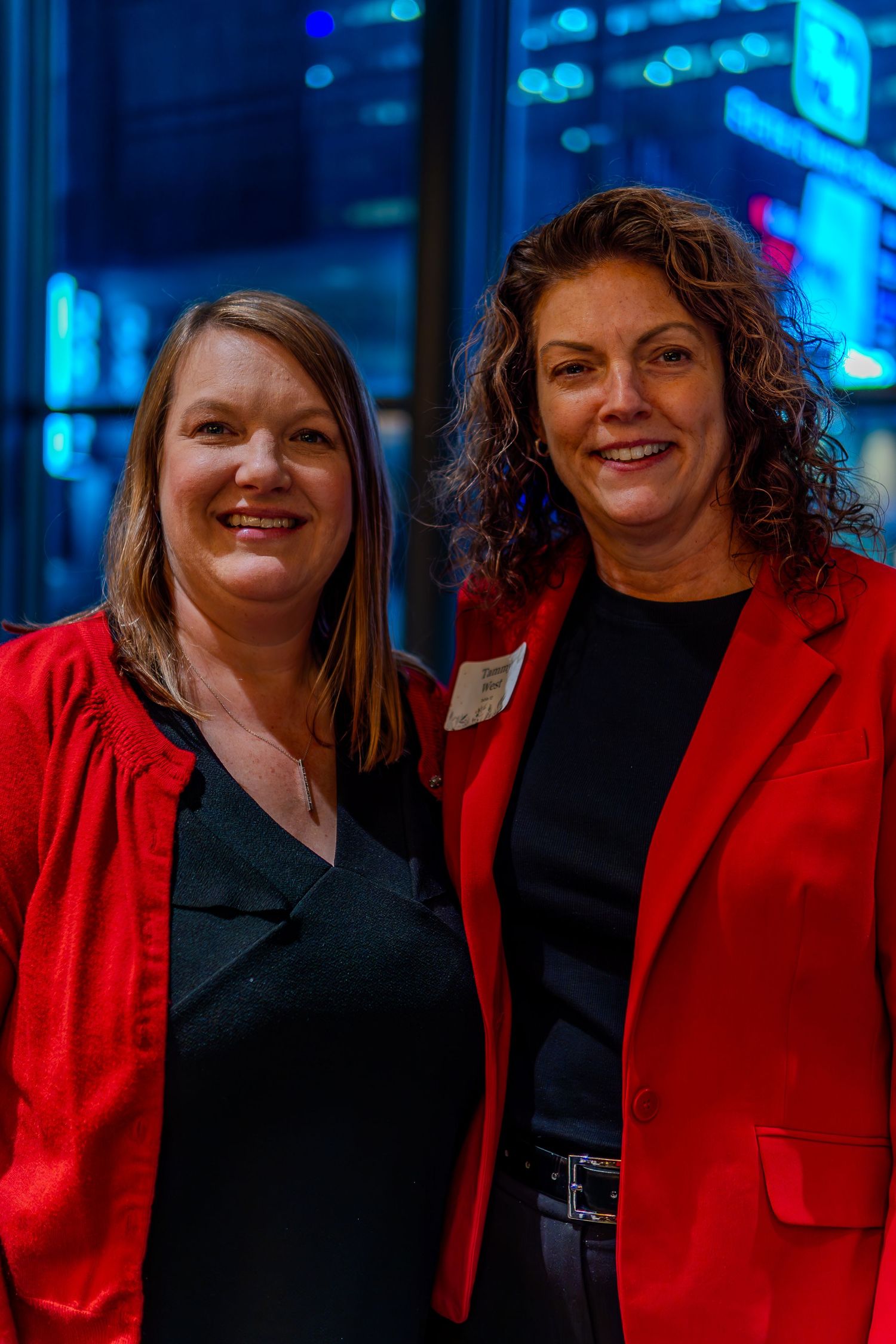 Two women in red blazers smile together at a formal evening event with city lights in the background.