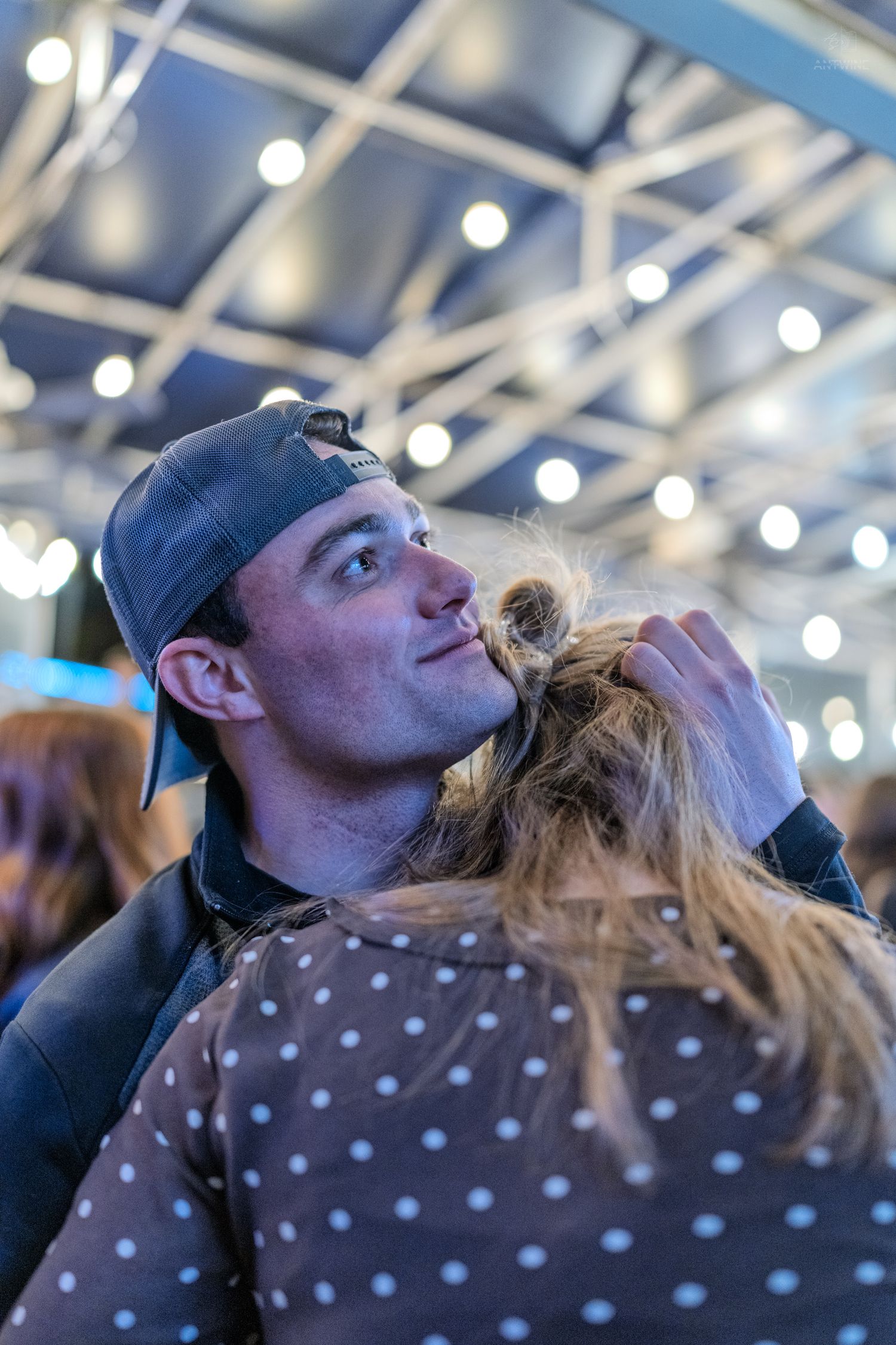 Man in baseball cap kissing the top of a womans head at an indoor event with string lights.