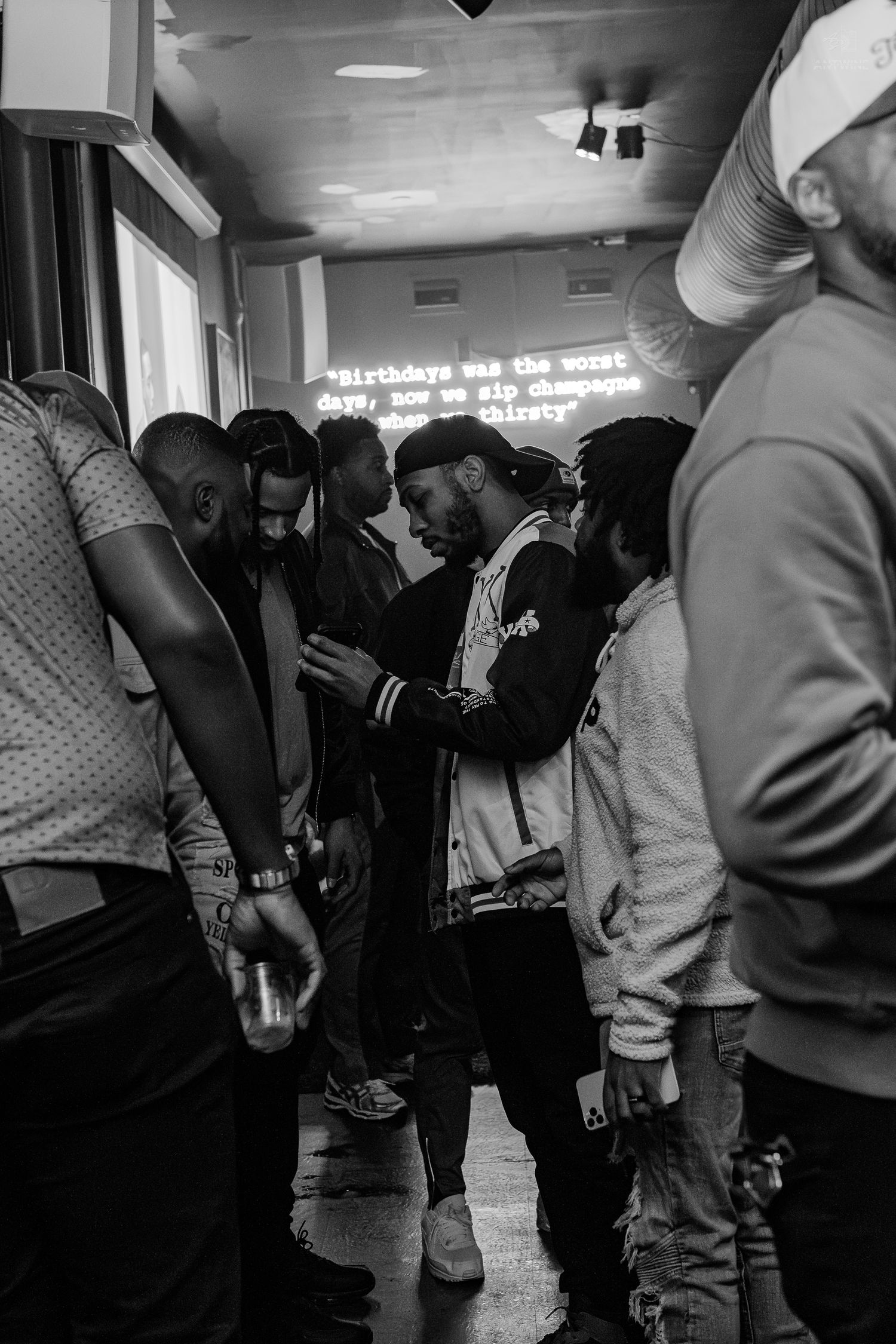 Black and white photo of a group of men gathered at an indoor party with neon sign in background.