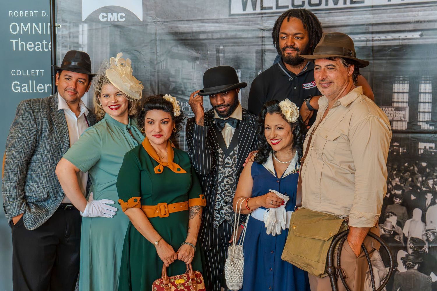 Group of seven people in vintage 1920s costumes posing together outside a theater gallery event.