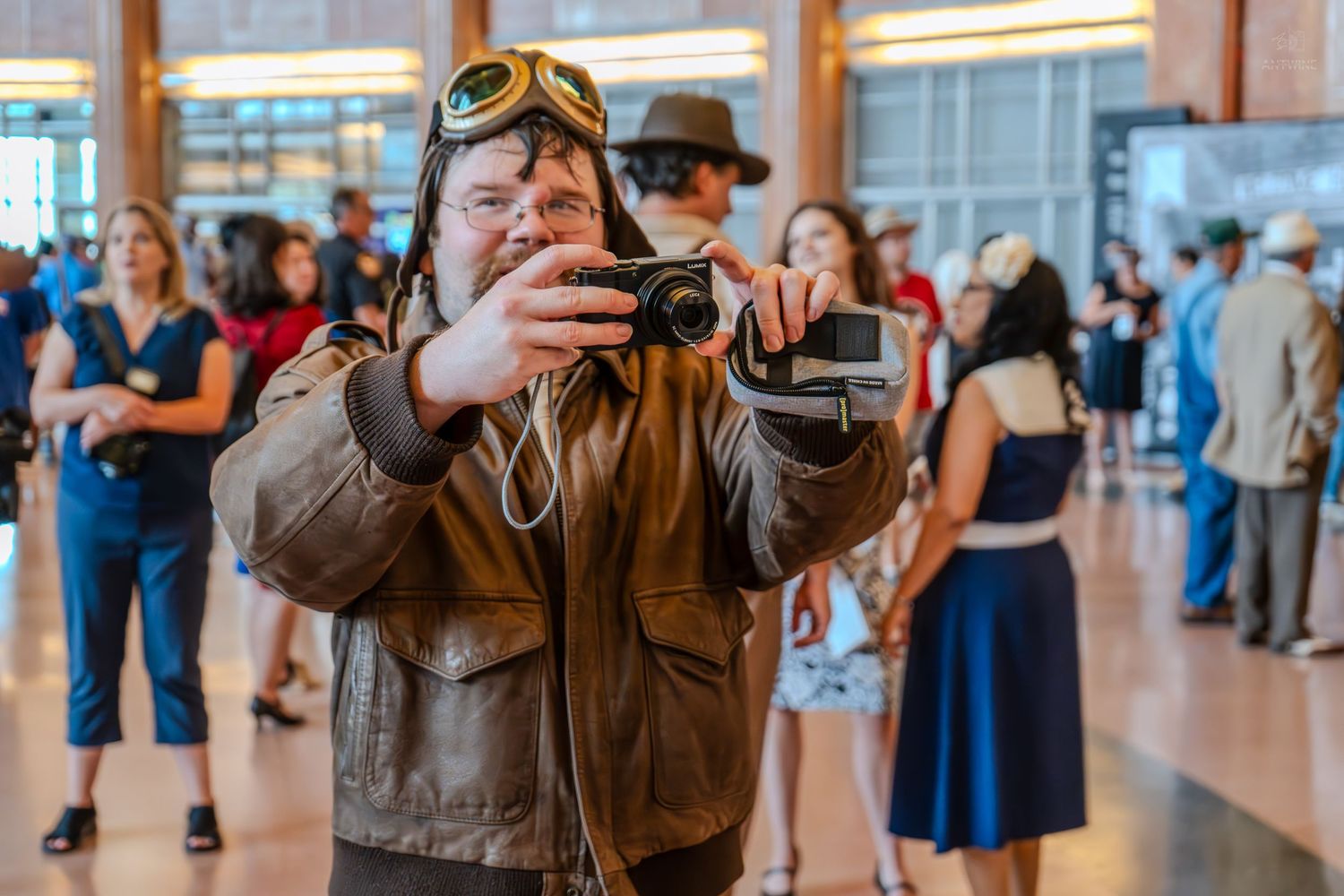 Man in vintage aviator jacket and goggles photographs with camera indoors at busy event.