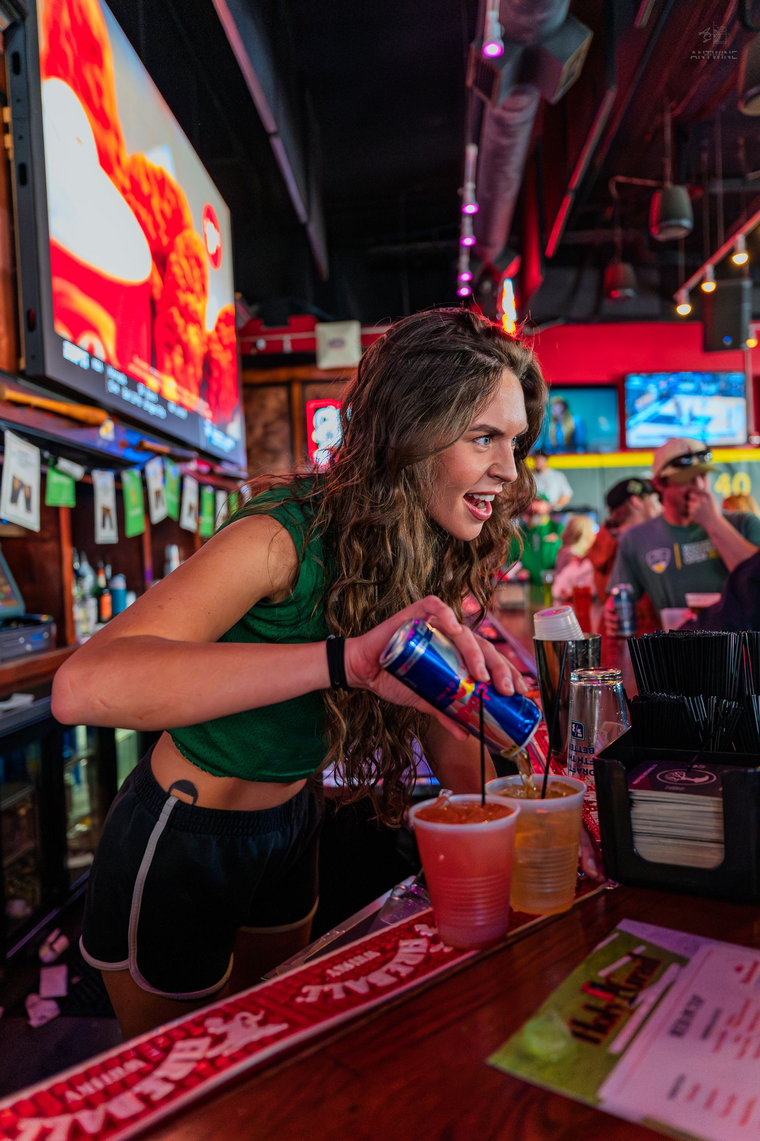 Smiling female bartender pouring a Red Bull energy drink into a cup at a lively sports bar with TVs in background.