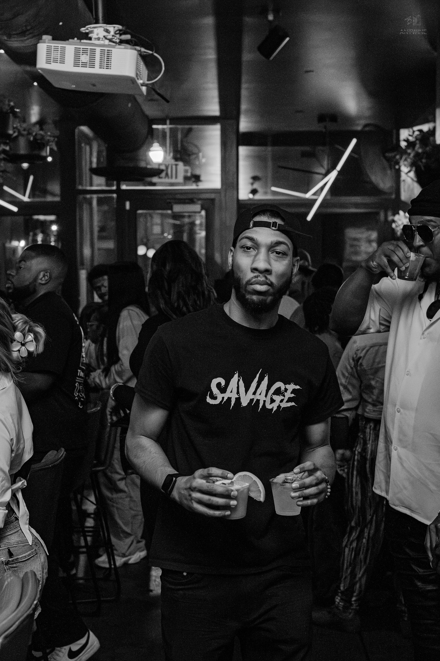 Black and white photo of a man in a Savage t-shirt holding a drink at a crowded bar event.