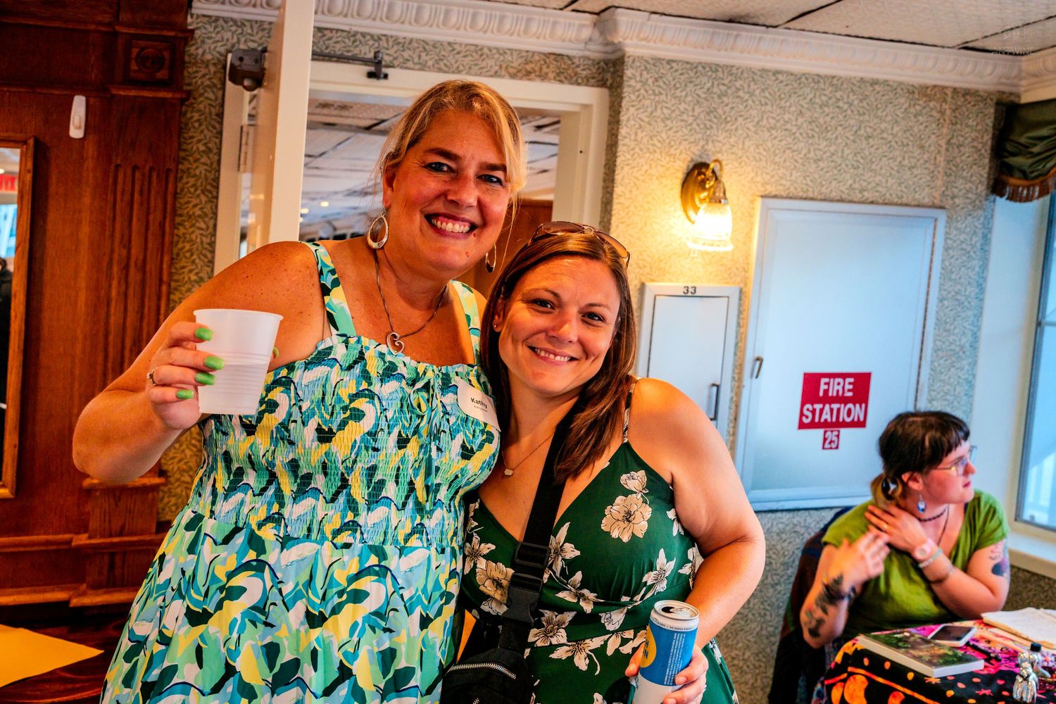 Two smiling women in floral dresses pose together at a casual indoor social gathering, one holding a cup.