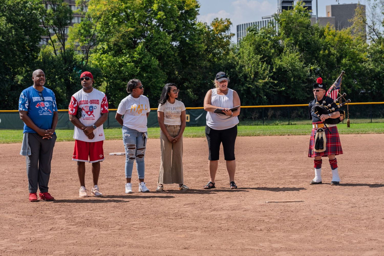 Group of people standing on a baseball field with a bagpiper in a red kilt during a ceremony.