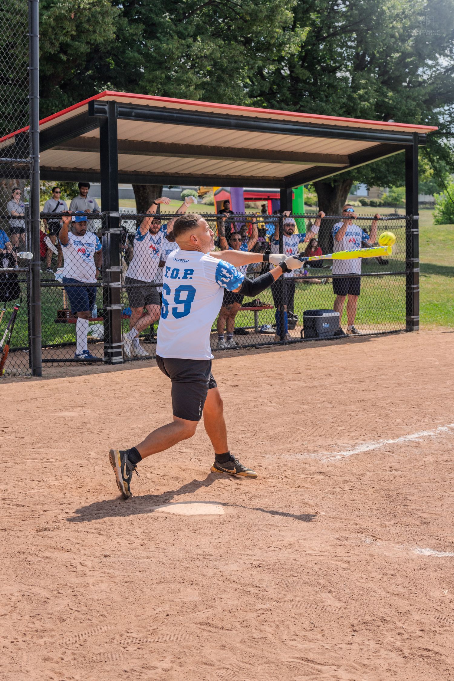 Player in jersey number 69 swings a bat at a softball game with spectators watching from behind a fence.