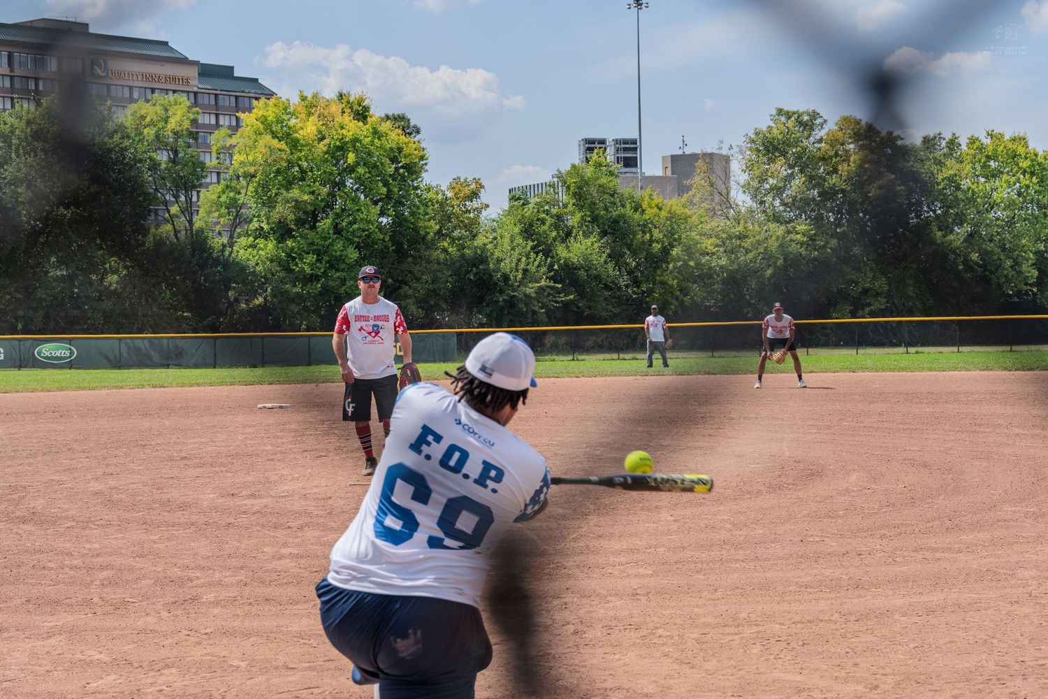 Softball catcher crouching to receive a pitch during an outdoor game viewed through a chain-link fence.