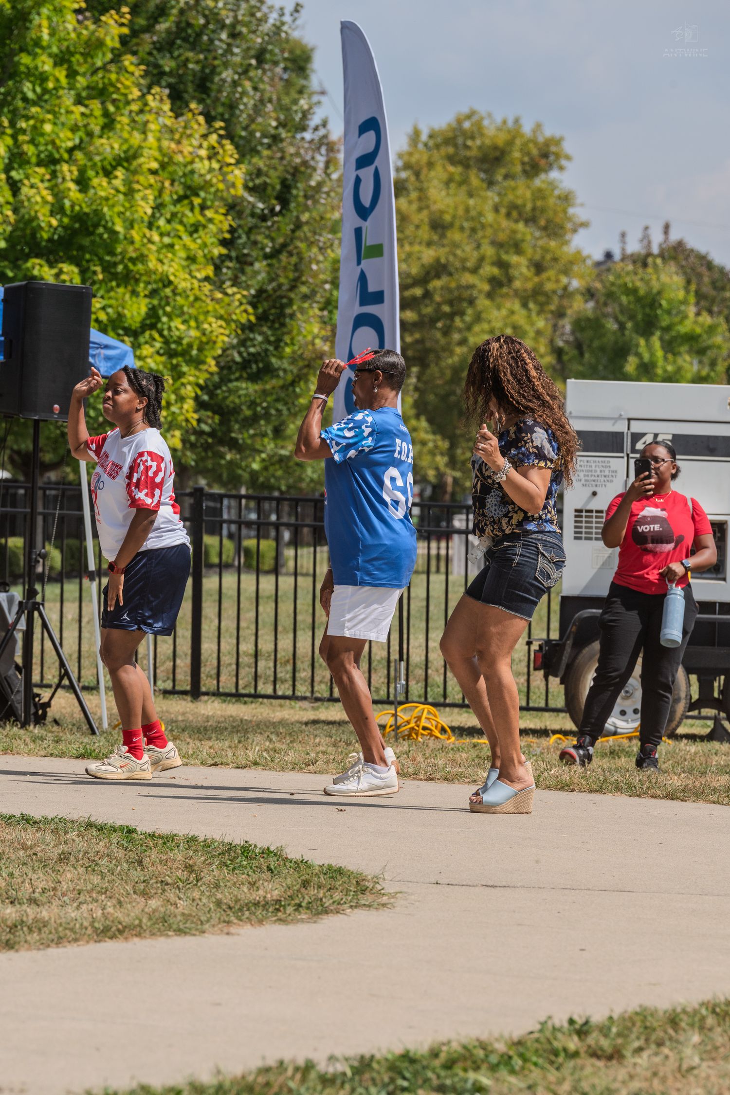 Group of people dancing outdoors at a sunny community festival near a tall blue banner.