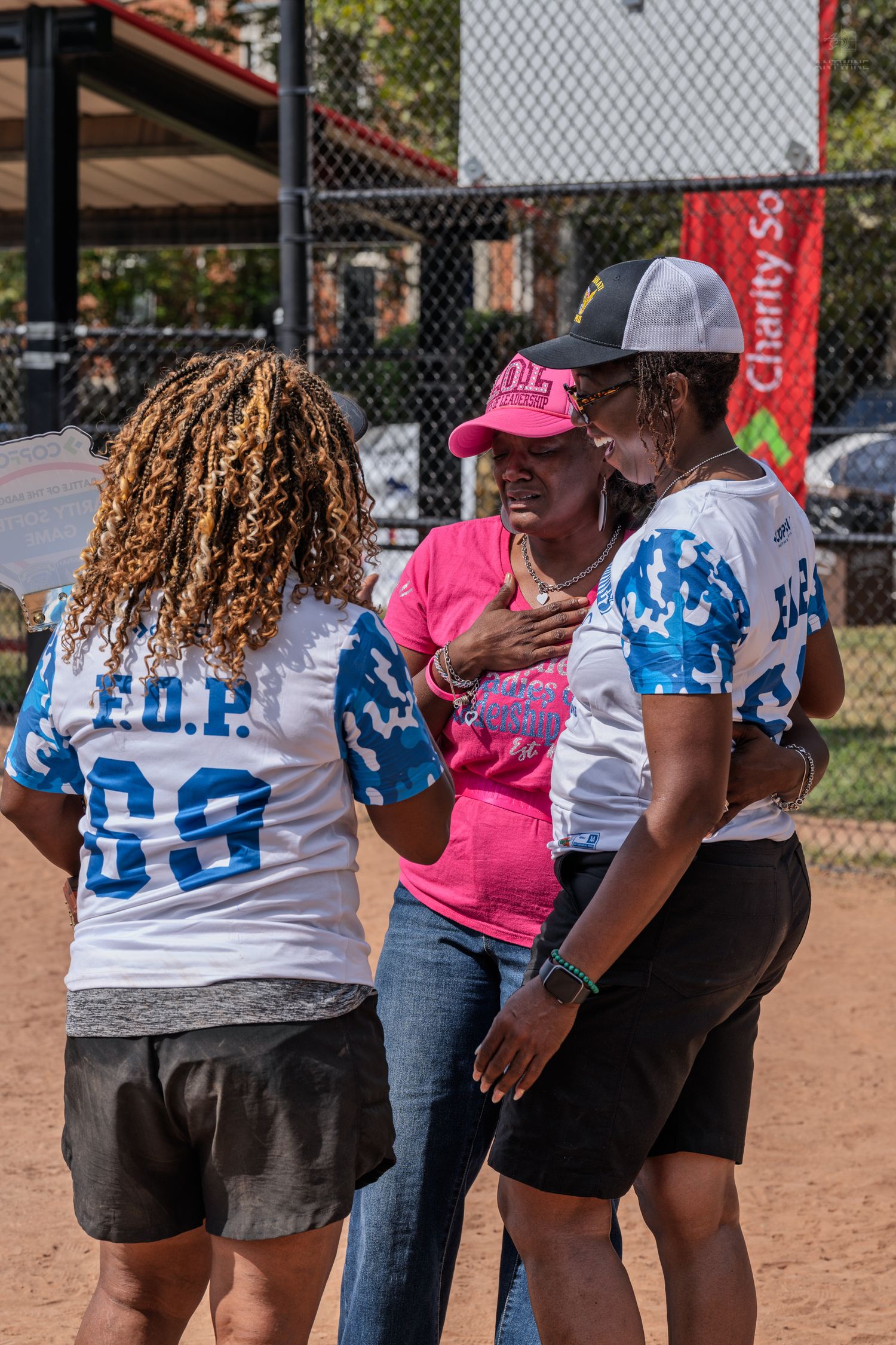 Three women in F.O.P. softball jerseys share a warm moment on a baseball diamond at a charity sports event.