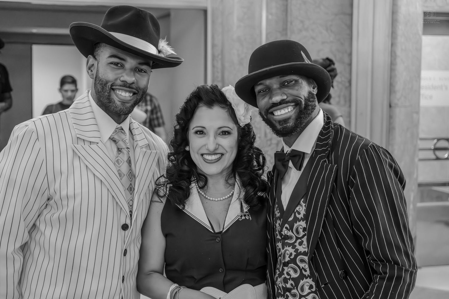 Three smiling people in vintage pinstripe suits and hats pose together in a stylish black and white photo.