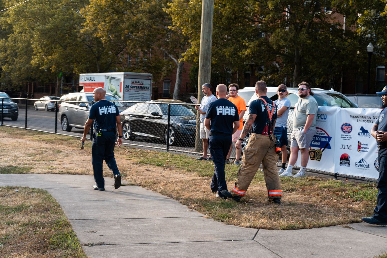 Firefighters in uniform walk along a sidewalk near a banner at an outdoor charity event.