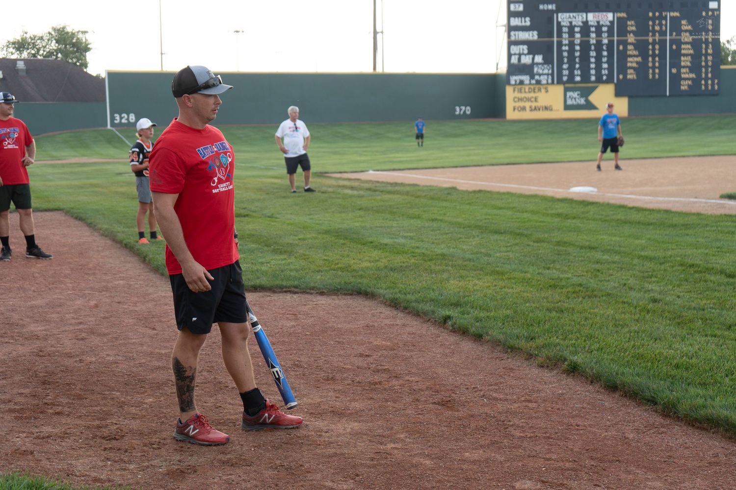 Adult softball player in red shirt stands with bat on field during evening recreational baseball game.