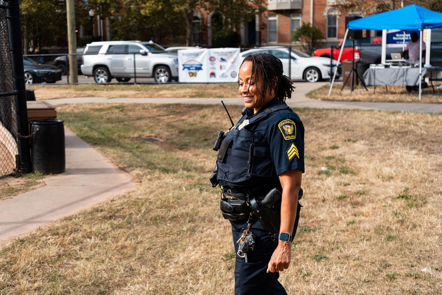 Female police sergeant smiling outdoors at a community event, wearing tactical vest and radio on a sunny day.