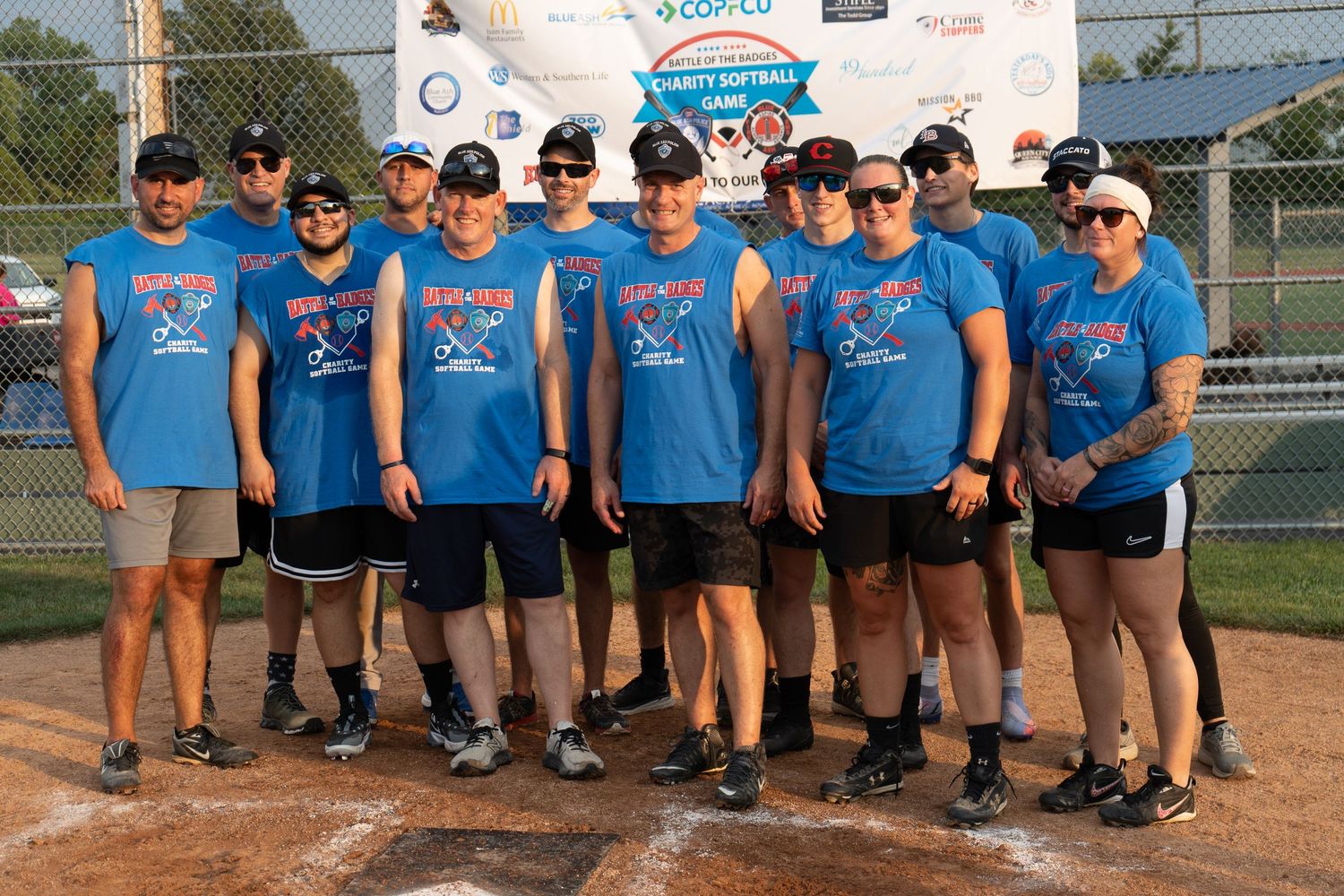 A charity softball team posing together in matching blue jerseys on a baseball diamond outdoors.