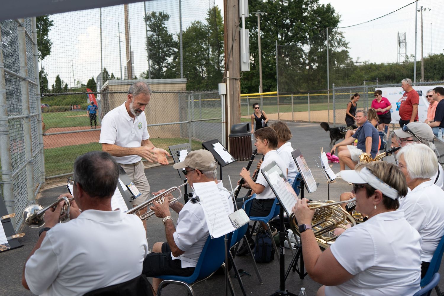 Outdoor brass band performance with conductor leading musicians in white shirts reading sheet music at a park venue.