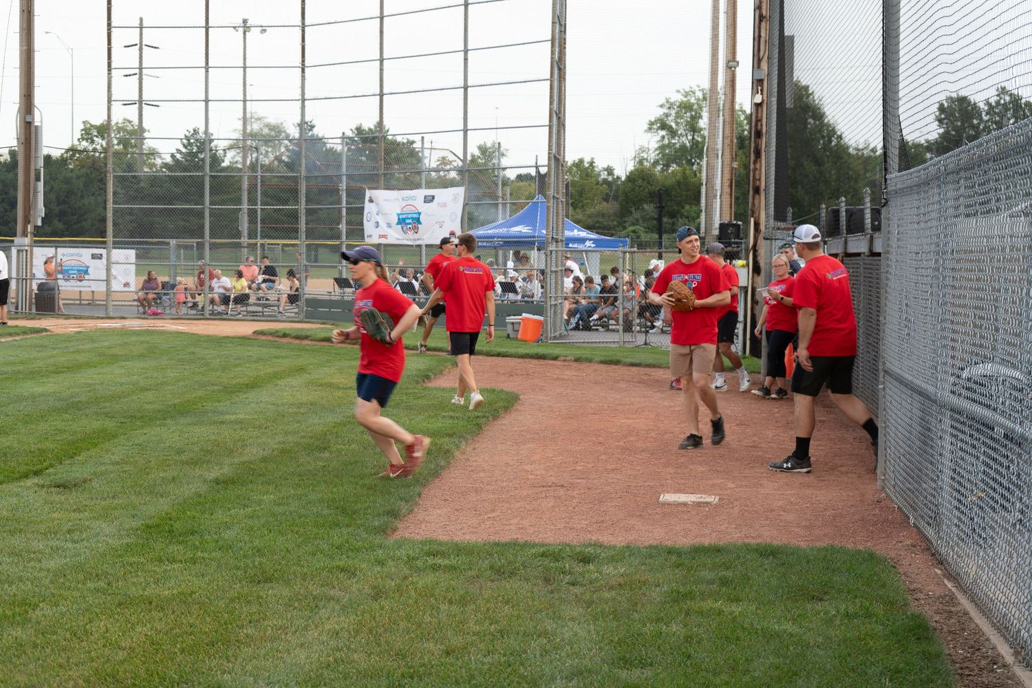 Adults in red shirts play softball on a green field during an outdoor recreational league game on a summer evening.