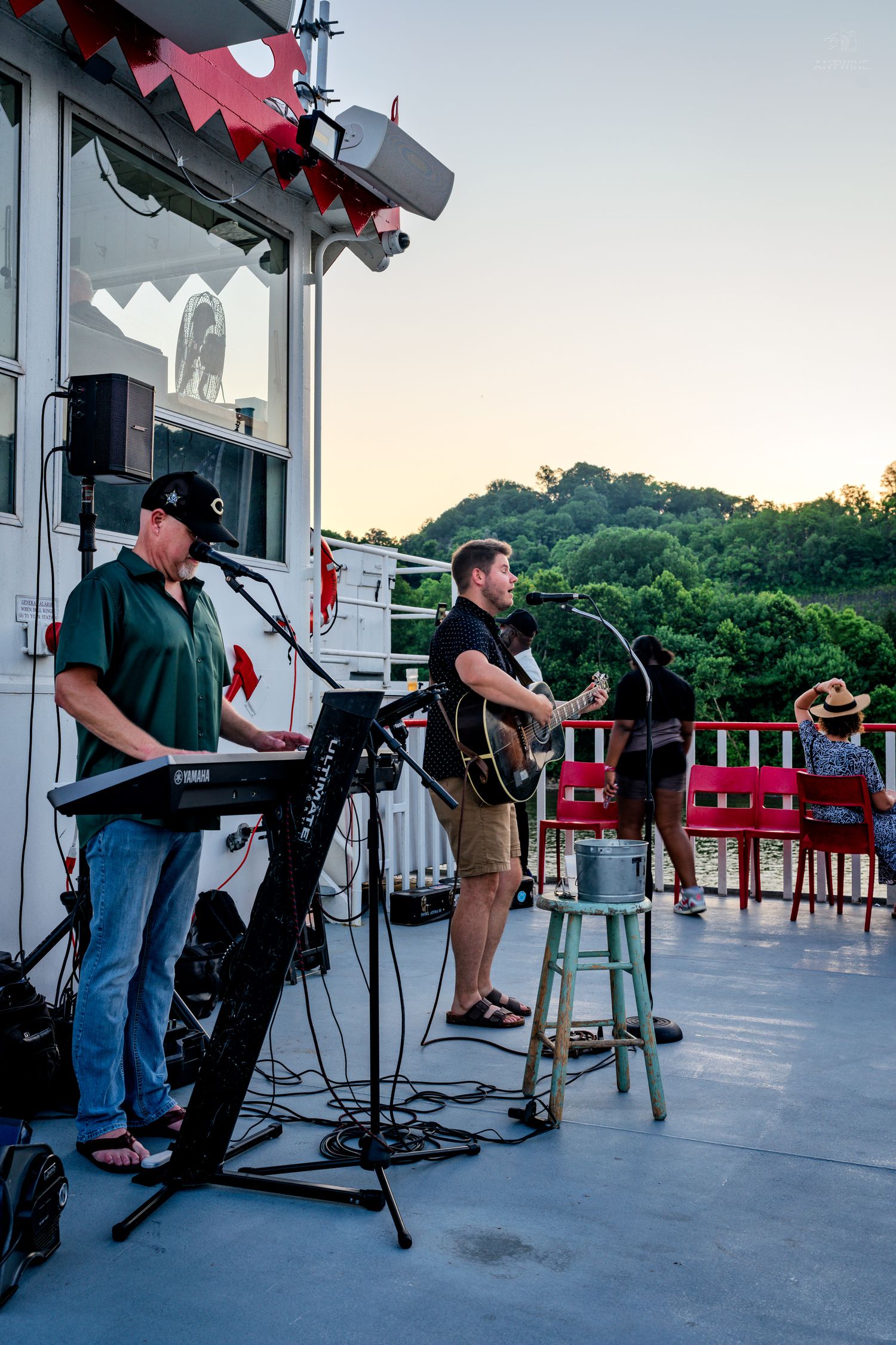 Live band performing on an outdoor deck at sunset, featuring a keyboardist and guitarist with scenic tree views.