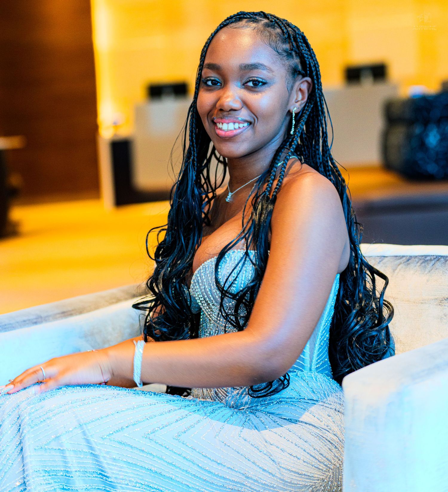 Young woman with long box braids smiling in a silver dress, seated in a modern lounge setting.