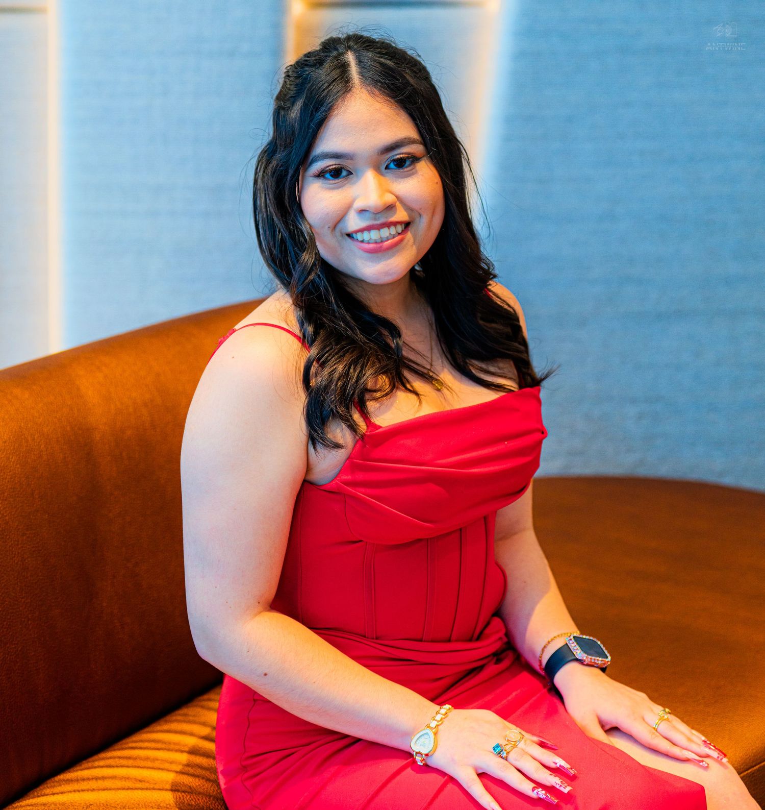 Smiling woman in a red off-shoulder dress seated on a brown leather sofa in an elegant venue.