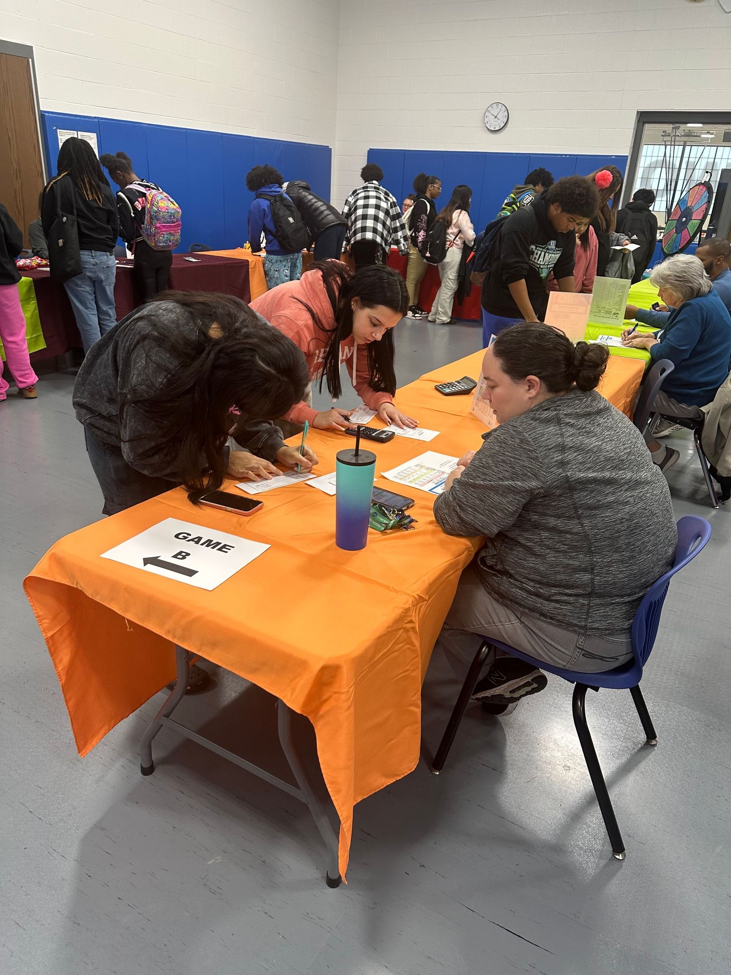 Students fill out forms at an orange-draped Game B table during a busy school gymnasium event.